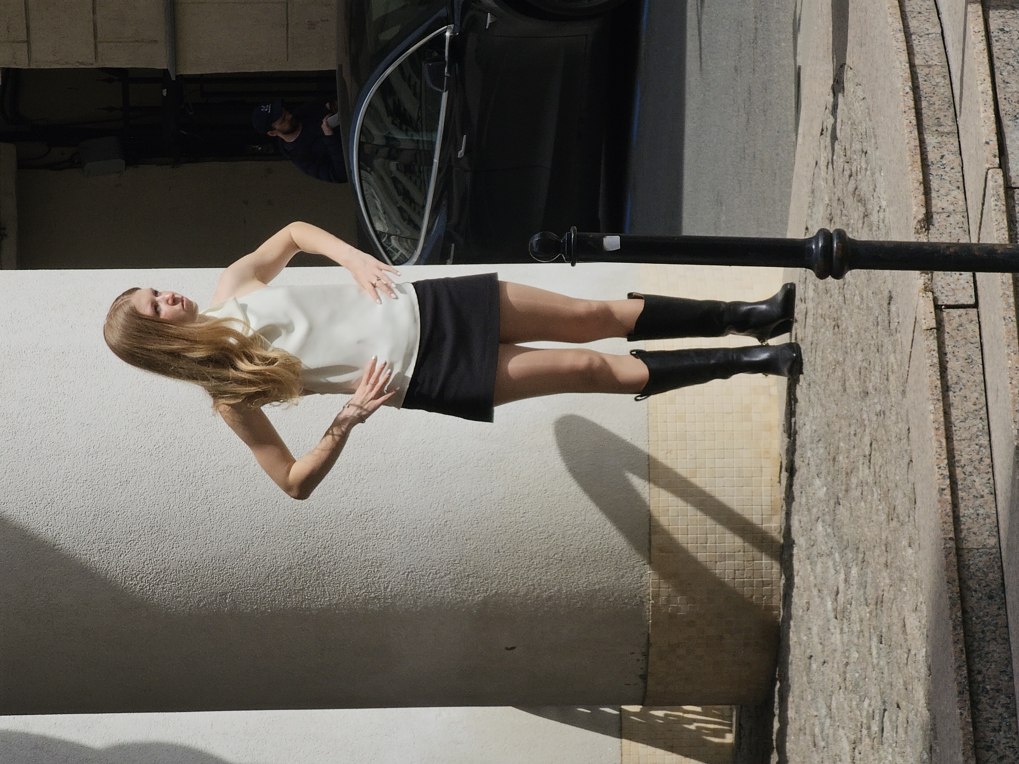 A fair-skinned woman in a white sleeveless blouse, black skirt, and black knee-high boots stands next to a black post in what appears to be an urban outdoor environment. The woman has long, light brown hair.
Behind her is a textured white wall. To the right of the woman is a pavement area with a pattern of stones and tile. Behind the woman and to her left is the side of a black car and a man wearing a dark baseball cap. The location is Moscow, Russia.