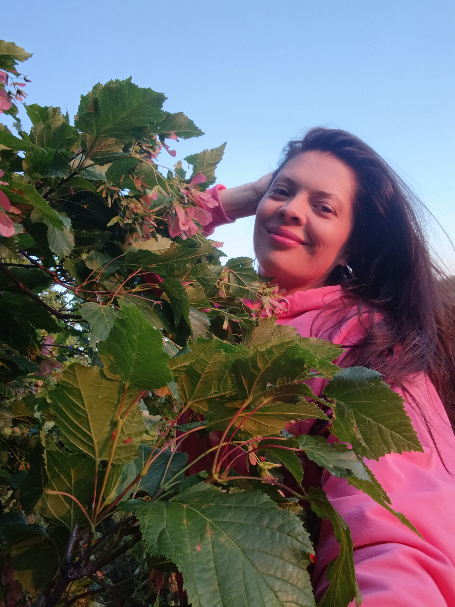 A medium shot shows a woman smiling and looking at the camera from behind a cluster of green leaves and some pink flowers. She is wearing a pink hooded sweatshirt and has her right hand resting on the back of her head. Her dark hair is loose and long. The background is a clear blue sky, suggesting an outdoor setting. The image quality is good, with clear focus on the woman and the foliage. There are no identifiable landmarks or contextual clues that indicate a specific location within Saratov, Russia.