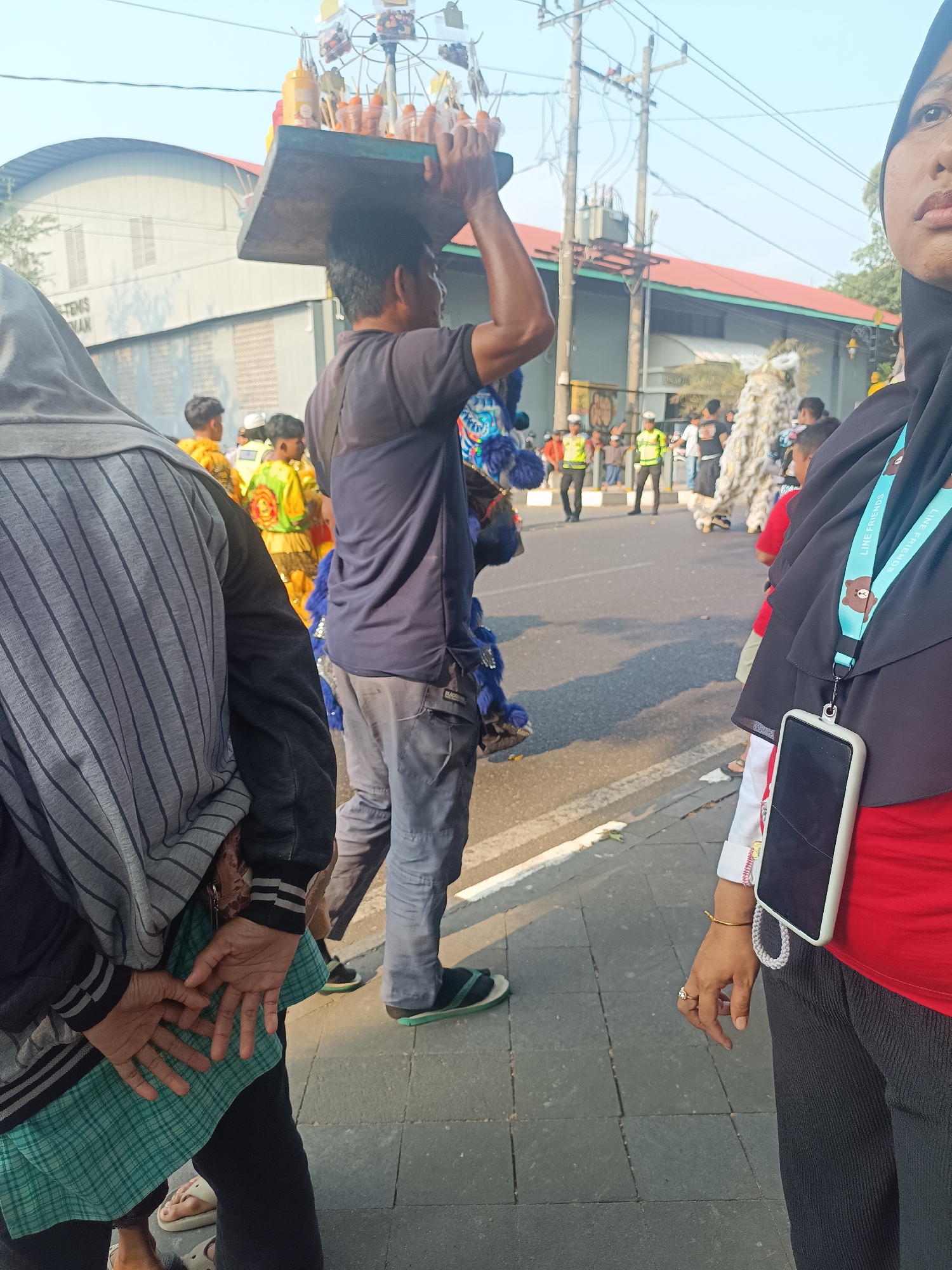 A street scene in Sleman, Indonesia, captures a lively gathering or parade. In the foreground, a person wearing a grey and black striped jacket and a green plaid skirt stands with their hands clasped behind their back. To their right, a woman wearing a black hijab and a red shirt is visible, with a mobile phone attached to her hijab by a lanyard. She wears a ring on her right hand. In the middle ground, a man carries a wooden board laden with various items, possibly food or goods, balanced on his head. He wears a dark blue t-shirt and grey pants. Behind him, individuals in colorful costumes, including one resembling a lion dance outfit and another with blue fringed embellishments, are present. Several other people, some wearing bright yellow vests, appear to be part of the event. The background features a building with a curved, grey roof, utility poles, and trees, suggesting an outdoor urban setting. The ground is paved with grey tiles. The overall atmosphere suggests a festive event taking place during daylight hours.