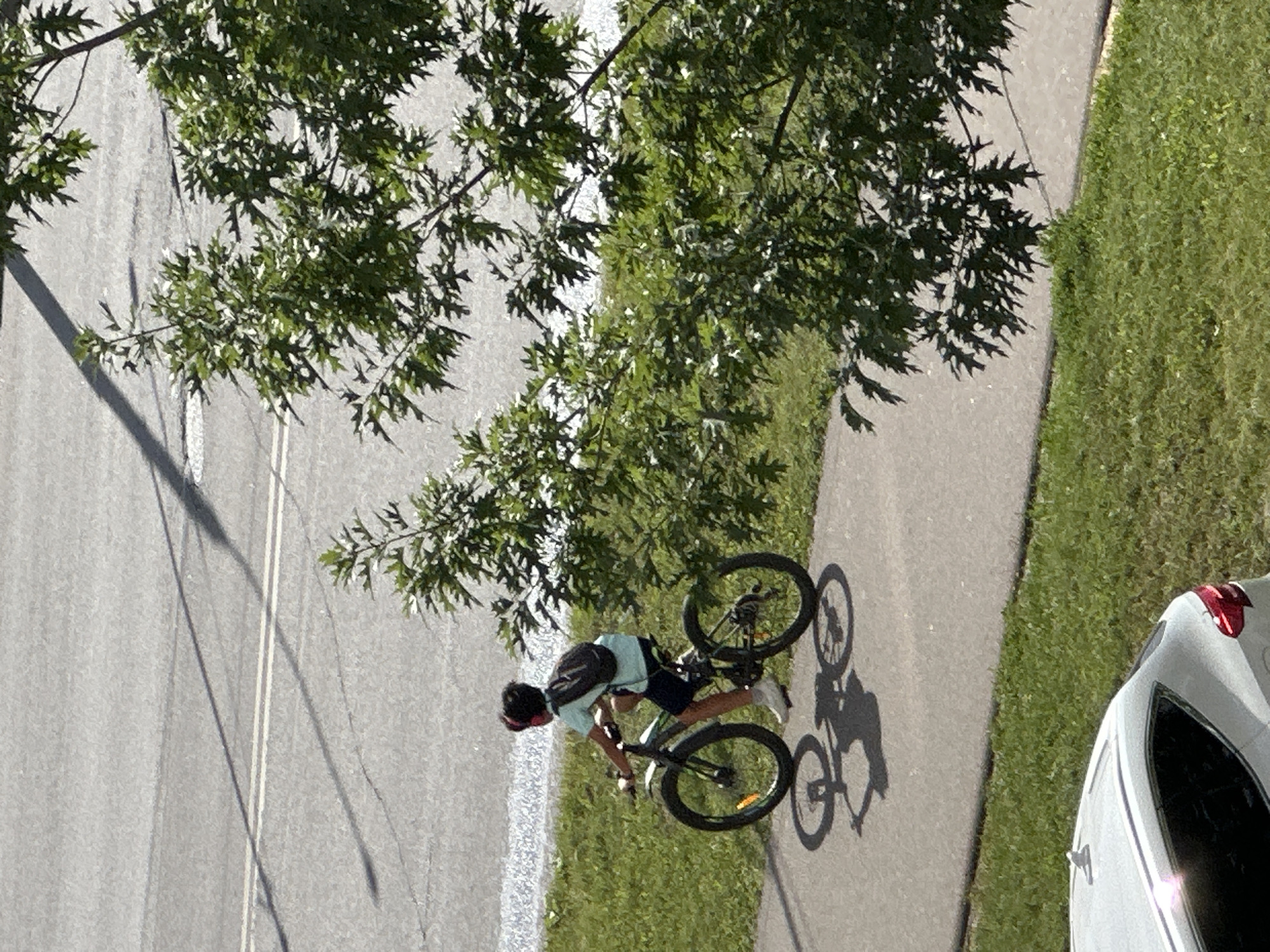In Podolsk, Russia, on a paved path adjacent to a grassy area and a road, a person rides a bicycle. The individual, wearing a light blue t-shirt, dark shorts, red headphones, and a backpack, is positioned on the right side of the path. The bicycle is dark-colored with thick tires. The sun casts distinct shadows of the bicycle and its rider onto the path. A portion of a white car is visible in the lower left corner, parked on the grass. Lush green foliage from a tree partially obscures the upper right portion of the scene. The road surface is asphalt with visible white lines, suggesting it is a street.