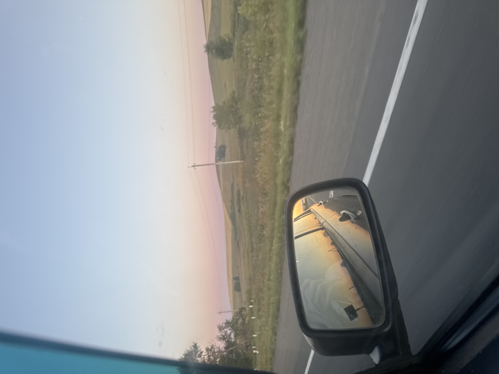 A side-view mirror of a car reflects a sunset over a rural landscape, as seen from a vehicle traveling on a road. The sky is a gradient of pale blue at the top, transitioning to soft pink and orange hues near the horizon. Rolling hills covered in green and yellow vegetation dominate the background, with scattered trees and utility poles visible. The road surface appears to be asphalt with a white line marking. The mirror also shows a portion of the car's interior, including what appears to be a white garment on the passenger seat, and the car's door handle. The image conveys a sense of movement and the beauty of nature during a sunset drive.