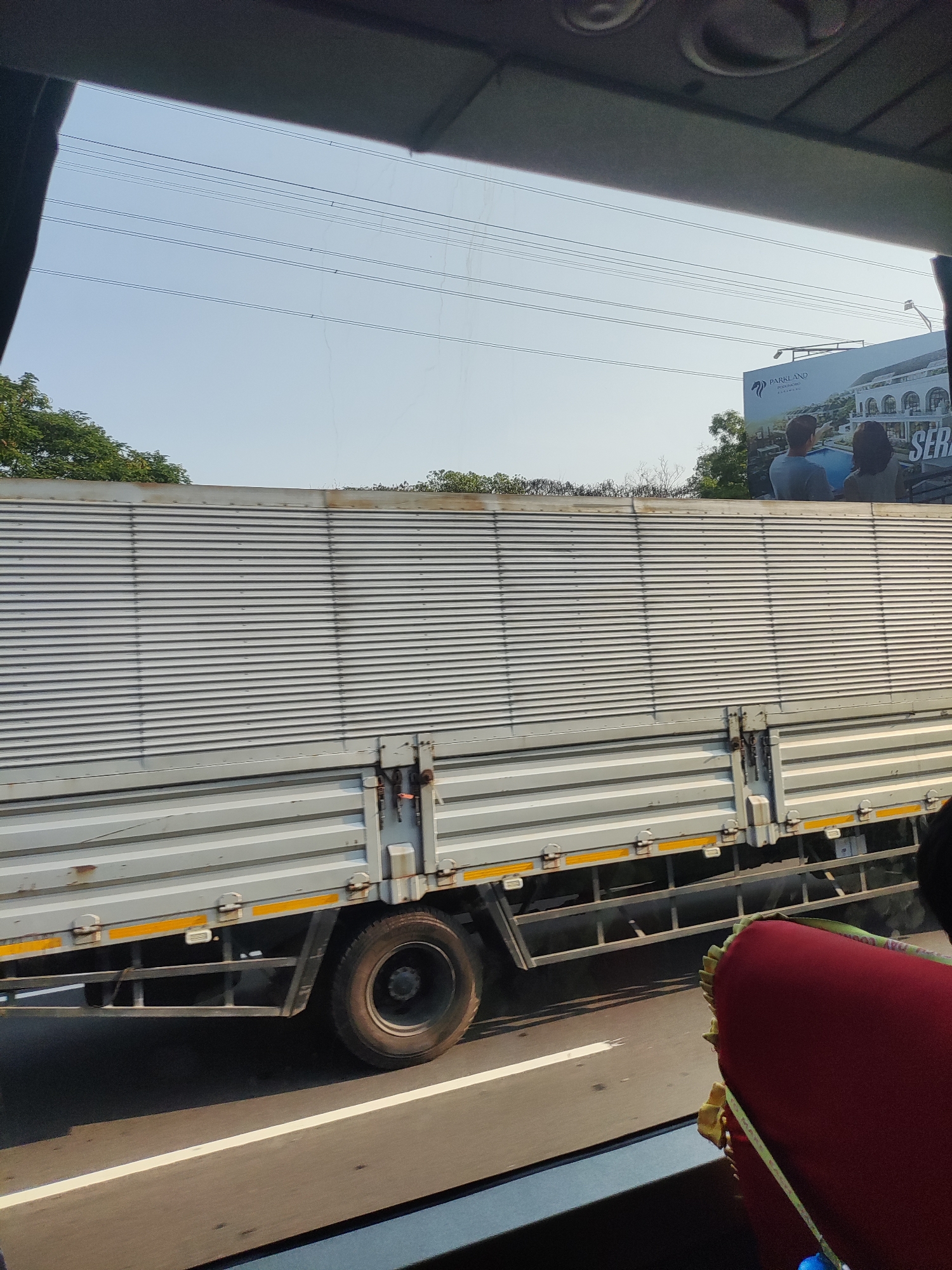The image captures a side view of a silver box truck on a highway under a clear blue sky. The truck's corrugated metal body is prominent, with yellow reflective stripes along its lower edge. Power lines are visible in the sky above. In the distance, a billboard displays an advertisement for "PARKLAND" housing and features an image of a building and two people. The truck's wheel and chassis are visible, as is the road surface with a white lane marker. The foreground shows a portion of what appears to be the interior of a vehicle, with a dark-colored edge along the bottom and a red, cushioned object with yellow straps. The scene is likely within the city of Karawang, Indonesia, given the context of the "PARKLAND" billboard, which is a real estate development in that region.