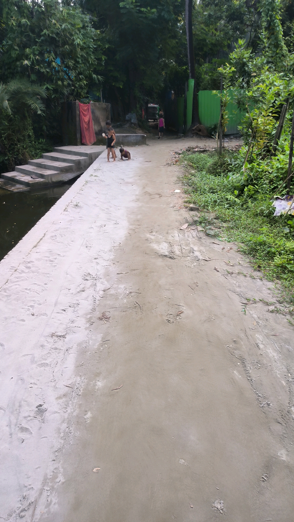 The image depicts a scene in Begumganj Upazila, Bangladesh. In the foreground, a sandy path runs diagonally from the bottom right towards the left, bordered by a concrete embankment and a body of water on the left. The embankment has a series of concrete steps leading down to the water. Several children are present near the embankment. One child in a black outfit stands near the steps, while another child is crouched down on the sandy ground. In the background, lush green foliage and trees dominate the scene. There are structures visible, including a red curtain hanging against a concrete wall, a bright green corrugated metal fence, and a glimpse of a tuk-tuk in the distance. The path appears to be unpaved and somewhat rough. The overall impression is of a rural or semi-urban environment with a natural landscape.