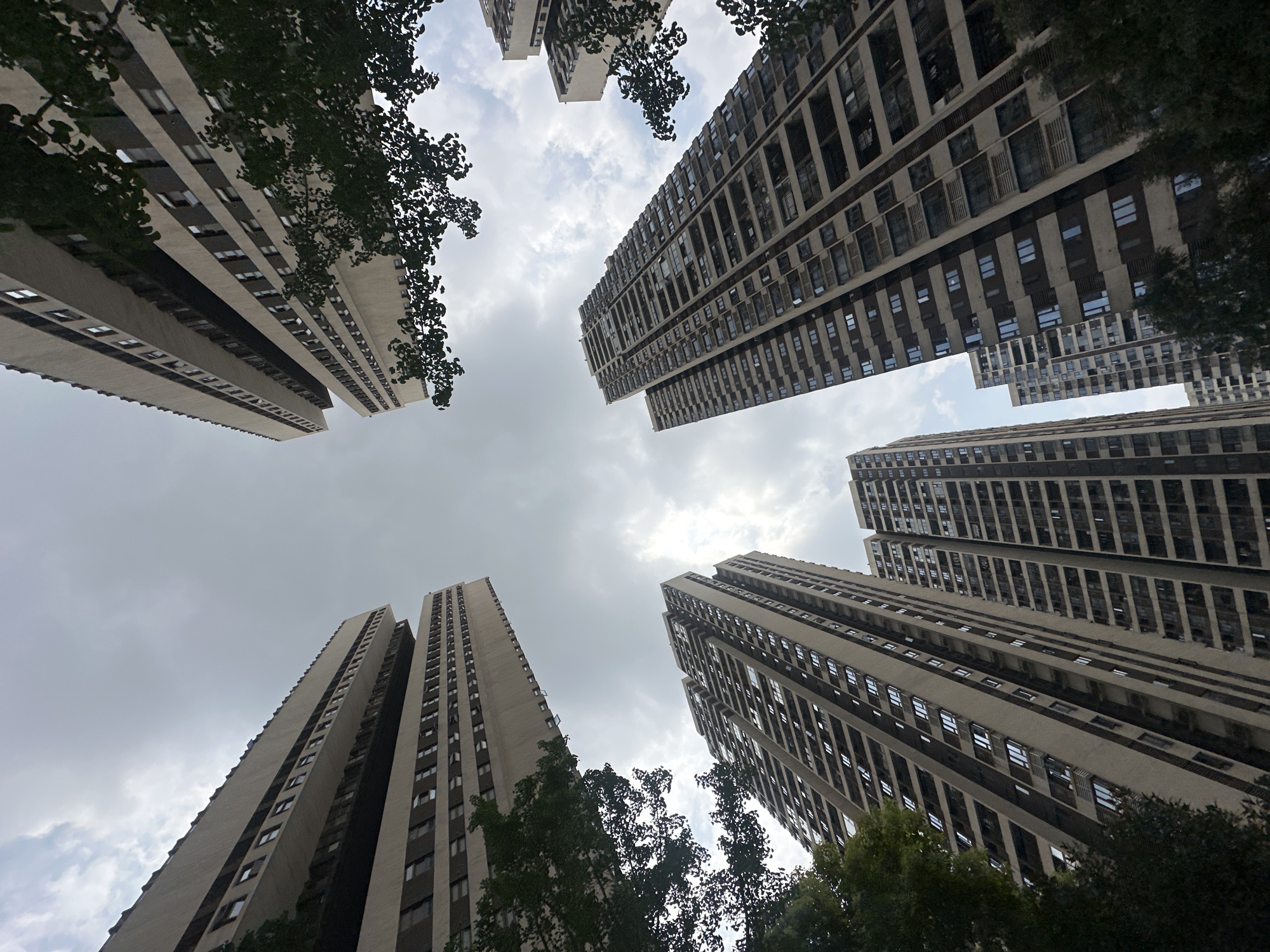 A low-angle shot in Chongqing High-tech Zone, China, reveals several tall apartment buildings, with a view pointing upwards towards the sky. The buildings are characterized by uniform rows of windows and a light-colored exterior. Some trees are present around the buildings, partially obscuring the view. The sky is overcast with grey and white clouds. There are no visible people or interactions in the frame.