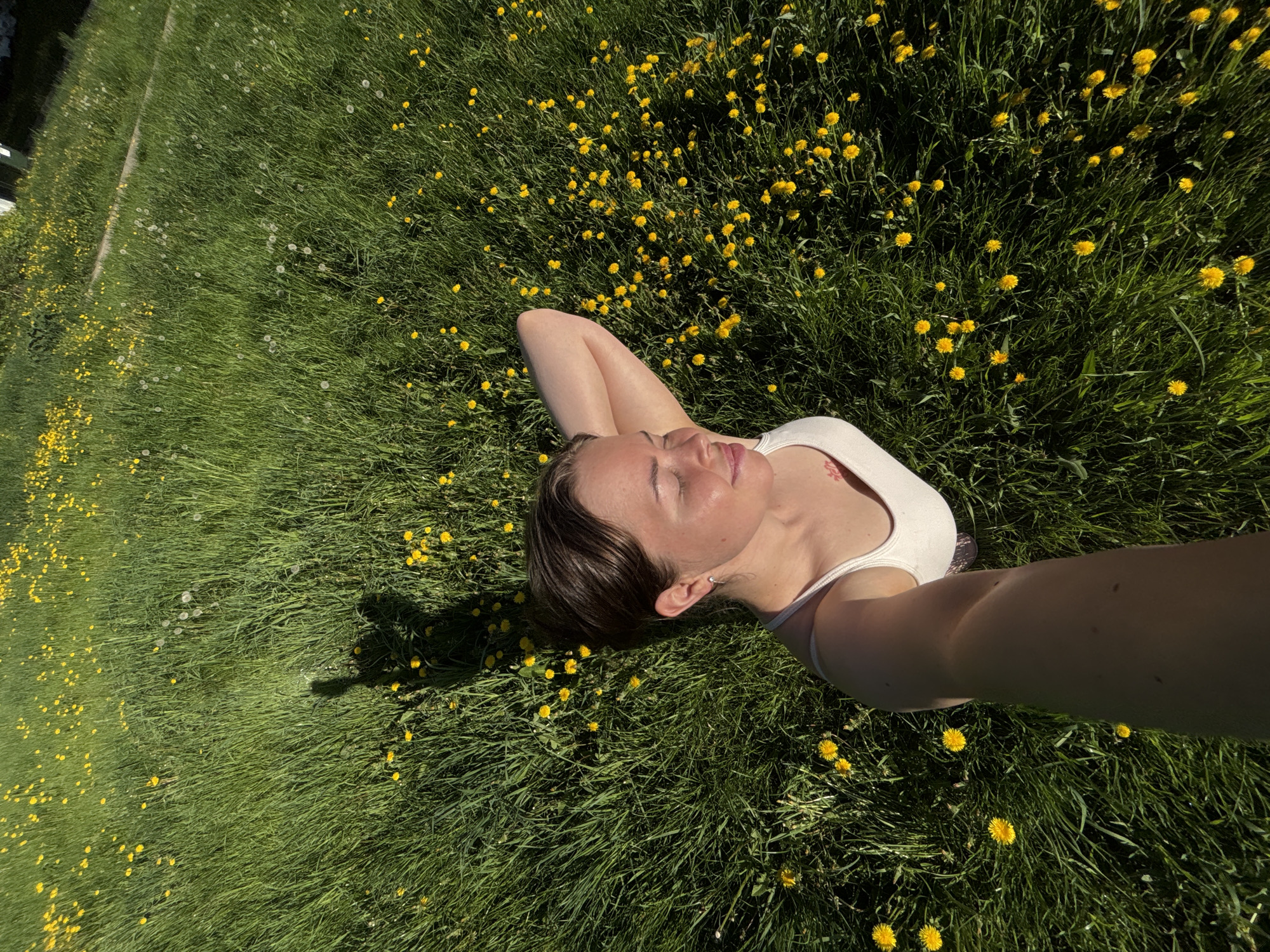 A woman is lying on her back in a field of grass and dandelions. She is wearing a white tank top and her arms are bent behind her head. Her eyes are closed and she appears to be relaxing. The photograph is taken from an overhead perspective, likely a selfie. The location cannot be confirmed as being in Izhevsk, Russia, based on the visual information provided.