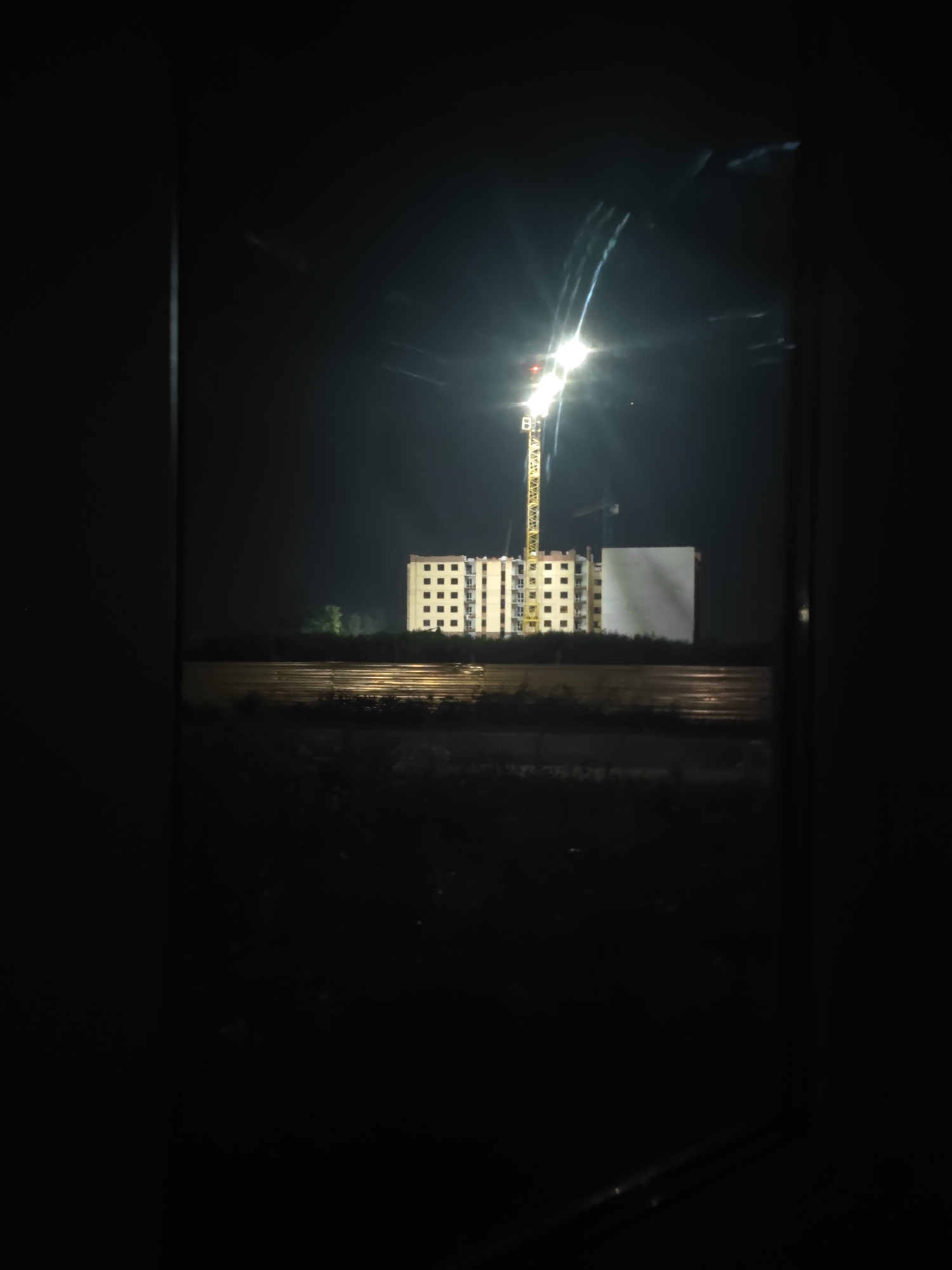 An outdoor, nighttime, low-angle medium shot captures a construction scene from within a dark frame. In the background, a tall, bright construction crane with a yellow lattice structure dominates the left side of the frame, with its powerful work light illuminating the scene. To the right of the crane, a partially constructed building with multiple floors and many windows is visible. The building is lit from within, creating a grid pattern of light and dark squares. In front of the building and crane, a flat, reflective surface, possibly wet ground or a body of water, mirrors the lights from above. Dark foliage lines the bottom of the frame. The foreground is filled with darkness, obscuring any details.