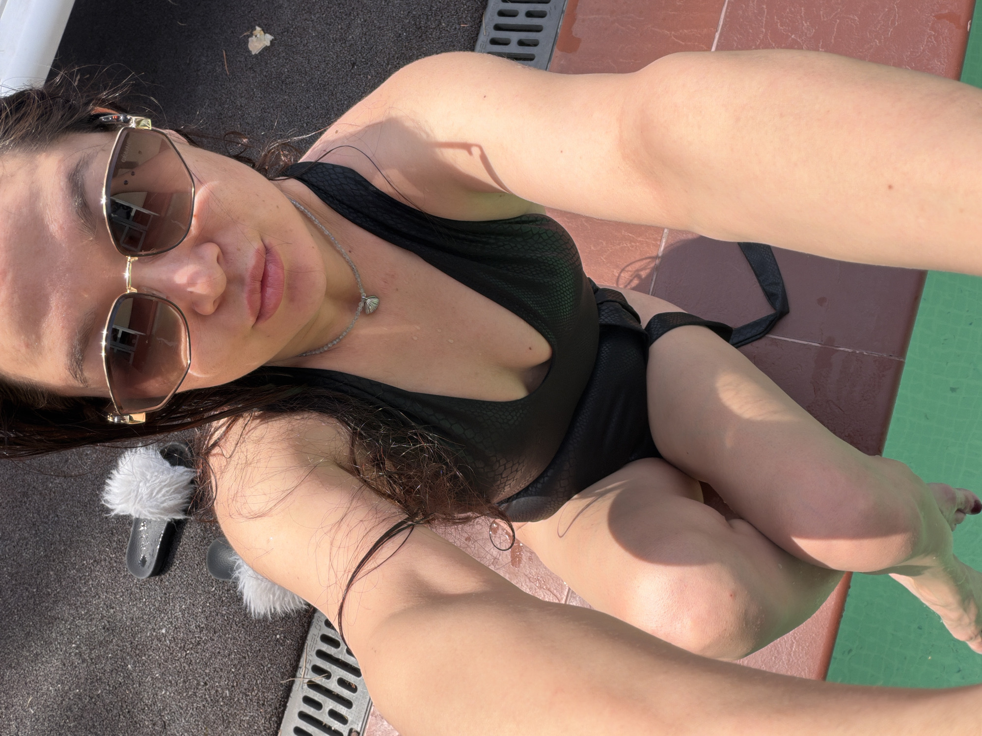 A woman wearing sunglasses and a black one-piece bathing suit is seated on the edge of a pool. She is wearing a necklace and has a pair of fuzzy sandals nearby. The location appears to be outdoors in Unknown, Unknown, next to a pool. The camera is held at an angle, giving a slightly distorted perspective of the scene.