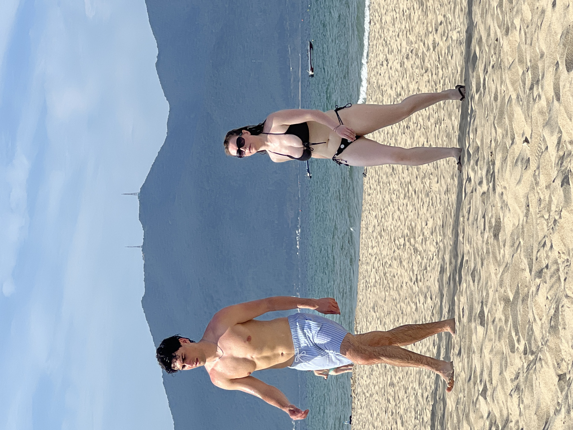 The image depicts a beach scene in Da Nang, Vietnam, with a clear sky and distant mountains. In the foreground, a man and a woman are walking on the sandy beach. The man, who is shirtless and wearing striped swim trunks, is on the left side of the frame. To his right, the woman is wearing a black bikini and sunglasses. The ocean is visible behind them, with a few boats in the distance. The overall atmosphere is one of a casual day at the beach.
