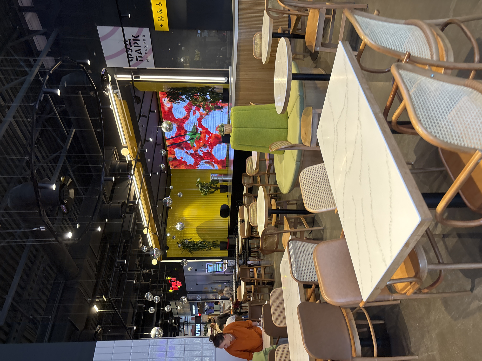This image captures a wide view of a modern, spacious indoor dining or food court area, oriented in a portrait style rotated 90 degrees counter-clockwise. The scene is well-lit by various artificial lights, including spotlights, strip lights, and decorative hanging spherical lights resembling disco balls.
On the right side of the frame, several dining tables and chairs are arranged. The tables have light-colored, possibly marble-patterned, rectangular and round tops. The chairs feature wooden frames with woven cane backs and seats, while others have brown leatherette upholstery. Some green upholstered sofas are visible in the mid-right.
Towards the middle-left, a large LED screen displays vibrant red and white abstract patterns, possibly depicting food or art. Below this screen is a bright yellow corrugated wall adorned with potted plants. The ceiling throughout the space is exposed, revealing dark industrial ductwork, beams, and numerous light fixtures.
In the bottom-center, a person with dark hair pulled back is seated at a table, wearing an orange or burnt orange long-sleeved top, looking downwards. Further to the bottom-left, other blurry figures can be seen in the distance, indicating a larger area with more activity.
Visible text includes a sign in the top-left corner that reads "ФУД ПАРК" (FOOD PARK) and "МЕРКУРИЙ" (MERCURY) on a white and pink background. Another yellow sign nearby shows an arrow pointing left with universal restroom symbols (male, female, disabled access). Towards the far bottom-left, a partially obscured sign appears to have Cyrillic text, possibly including "ПАРНИ" (GUYS), but it's largely illegible due to distance and blur. The overall setting suggests a vibrant and contemporary commercial dining environment.
