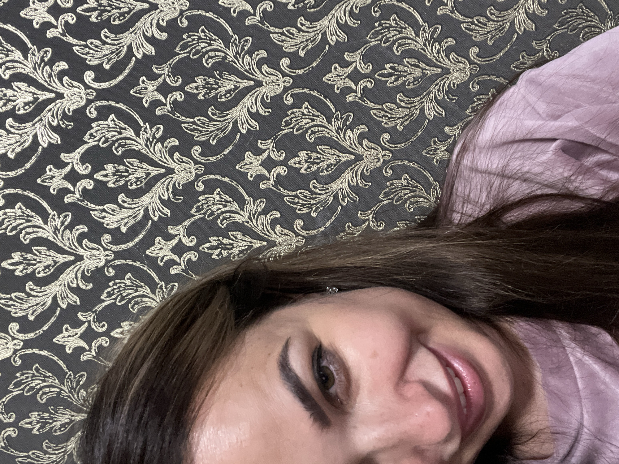 A close-up, waist-up shot captures a woman's face and upper torso against a dark gray damask wallpaper with a gold floral pattern. The woman, positioned on the left side of the frame, has dark brown hair that falls over her shoulder. Her visible eye is a warm brown, and her eyebrows are dark and well-defined. She is smiling, revealing her teeth and a hint of pink lipstick. She is wearing a soft pink, long-sleeved top. A small, clear stud earring is visible in her left ear. The lighting is soft, highlighting her features and the texture of the wallpaper. The scene appears to be indoors, in an environment with decorative wallpaper.