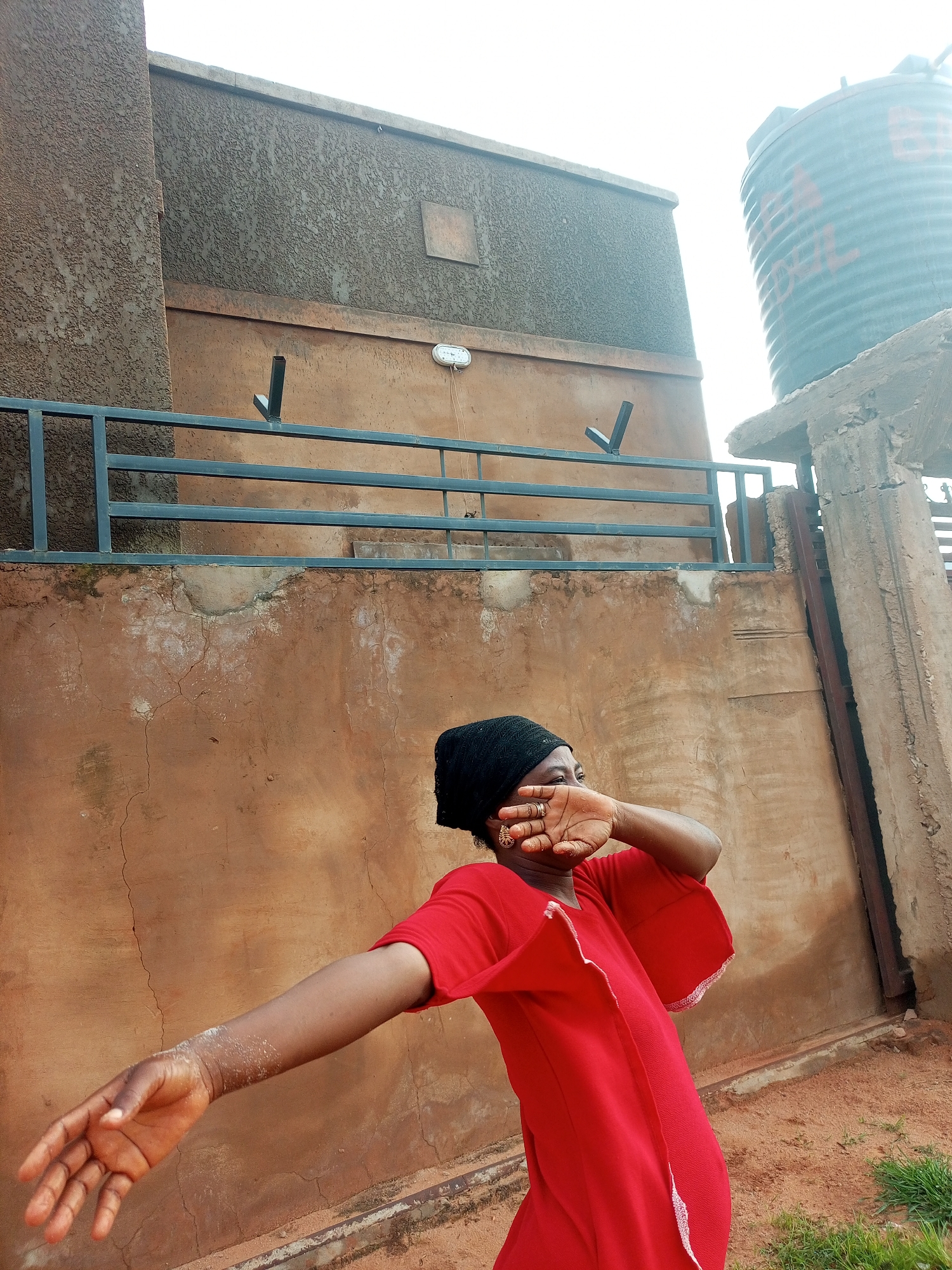 A woman wearing a red dress and a black headscarf stands in front of a brown textured wall. Her right hand is covering her face, and her left arm is extended to the side. She appears to be in an outdoor setting, possibly a residential area, given the presence of a water tank and a metal fence. The ground is covered with dirt and some patches of grass. The overall scene suggests a casual, everyday moment. The location is specified as Tempure, Nigeria.