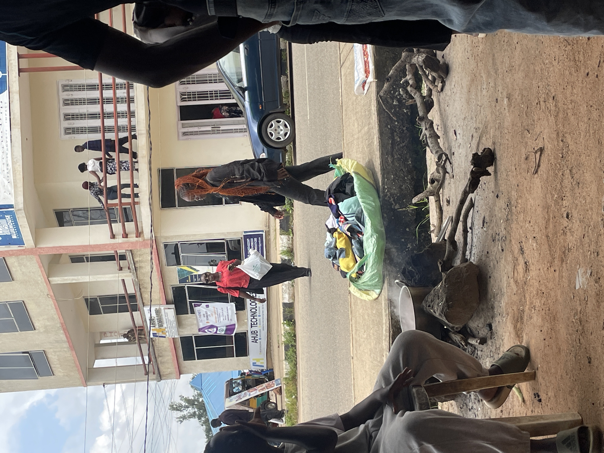 The image depicts a street scene in Jos, Nigeria, with a building in the background. In the foreground, a person is seated next to a fire with a pot, suggesting food preparation. Nearby, a pile of clothing is laid out on a green tarp.
In the middle ground, a woman with bright orange hair walks across the street, carrying a bag. She is dressed in dark clothing. On the sidewalk, another woman in a red shirt and dark pants walks away from the camera, also carrying a white plastic bag.
Further in the background, people are visible on a balcony of the building. The building has multiple stories with windows and balconies. A blue car is parked on the street. Signs are posted on the building, one of which appears to be advertising "AHUB TECHNOLOGIES." A small three-wheeled vehicle is also visible on the street. The overall environment appears to be a bustling urban area with signs of daily life and commerce.