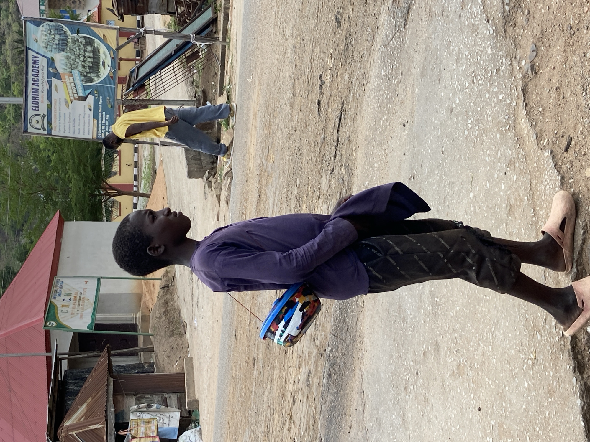 A young person in a purple shirt and dark shorts walks along a concrete road in Mararaba, Nigeria, carrying a colorful container hanging by a string. A person in a yellow shirt and grey pants walks away in the background. Signs for "CRCN" and "Elohim Academy" are visible, indicating the location.