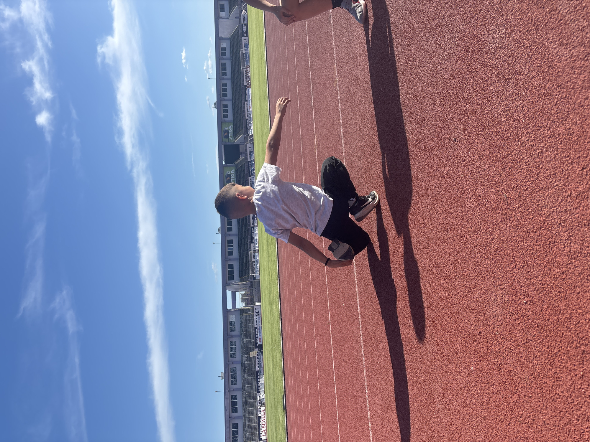 The image captures a bright, sunny day at an athletic stadium. A young boy, dressed in a white t-shirt and dark pants, is kneeling on the red running track. His right arm is raised, and he appears to be interacting with someone or something just out of frame, evidenced by the shadow of another person standing nearby. The stadium's stands, visible in the background, have advertisements displayed, including the brands WINBET and TIGA. A green grass field is located to the left of the running track. The sky is a clear blue with scattered white clouds. Based on the stadium architecture and the Winbet advertisements, it is likely the Ivaylo Stadium in Veliko Tarnovo, Bulgaria.