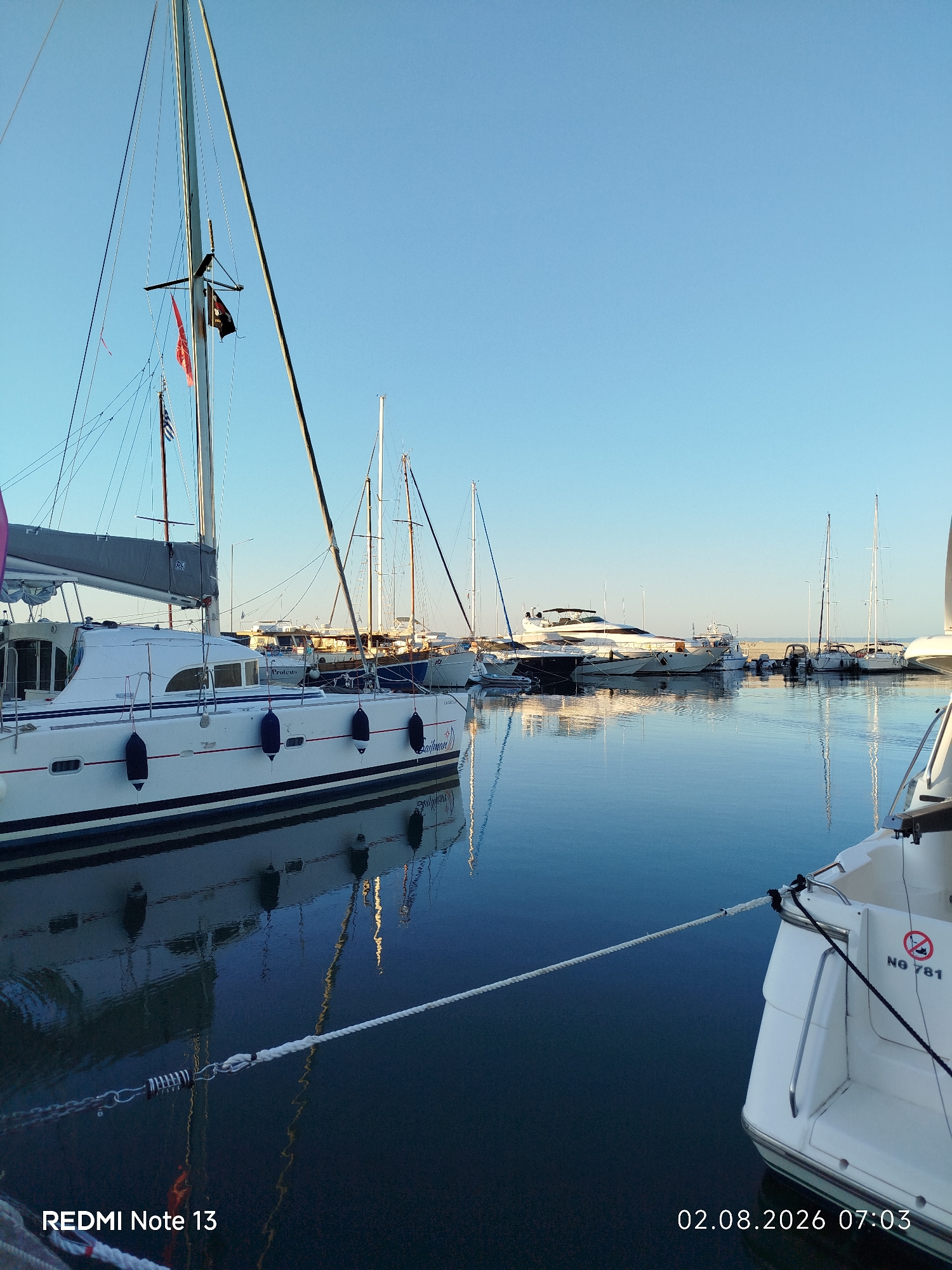A wide shot captures a marina in Sithonia Municipal Unit, Greece, under a clear blue sky. Several sailboats and yachts are docked, their masts creating a vertical pattern against the horizon. The water is calm and reflects the boats and the sky. In the foreground, the stern of a white boat with the registration "NG 781" is visible, secured by a rope. The timestamp "02.08.2026 07:03" and "REDMI Note 13" are superimposed on the image. The lighting suggests early morning.