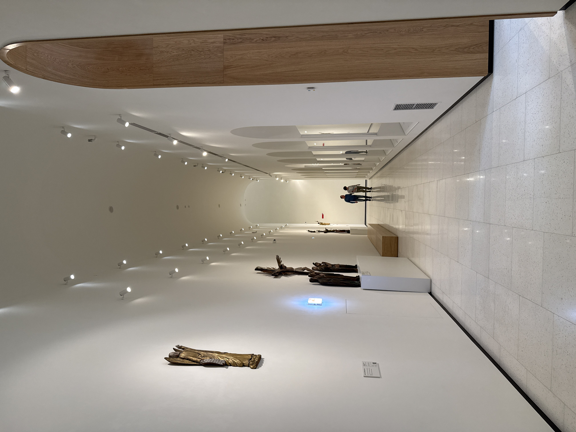 The image shows the interior of the Perm State Art Gallery, located in Perm, Russia. The gallery space is brightly lit with a white color scheme, with the floor tiled in a light, speckled pattern. A series of recessed lighting fixtures run along the upper part of the walls and ceiling. A large, wood-paneled structure is at the top left of the image.
The gallery features several sculptures displayed on the floor. The sculptures are primarily of crucified figures. There are two people in the distance, seemingly observing the art. The overall impression is of a minimalist and modern art space showcasing religious-themed artwork.