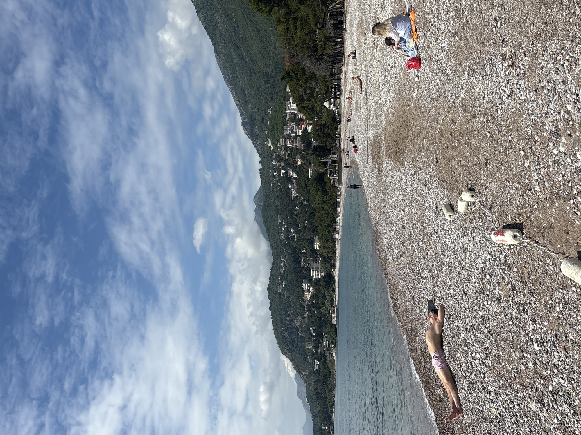 The image shows a beach on the coast of Bar, Montenegro. The beach is made of small, light-colored pebbles. A mountain covered in dense greenery with houses nestled along its slope rises along the beach. The sky above is blue with scattered white clouds. A man lies shirtless on the beach near the water, wearing pink swimming trunks. Further up the beach, a woman lies on a blanket with her head covered. Buoys float near the edge of the water, demarcating a swimming area.