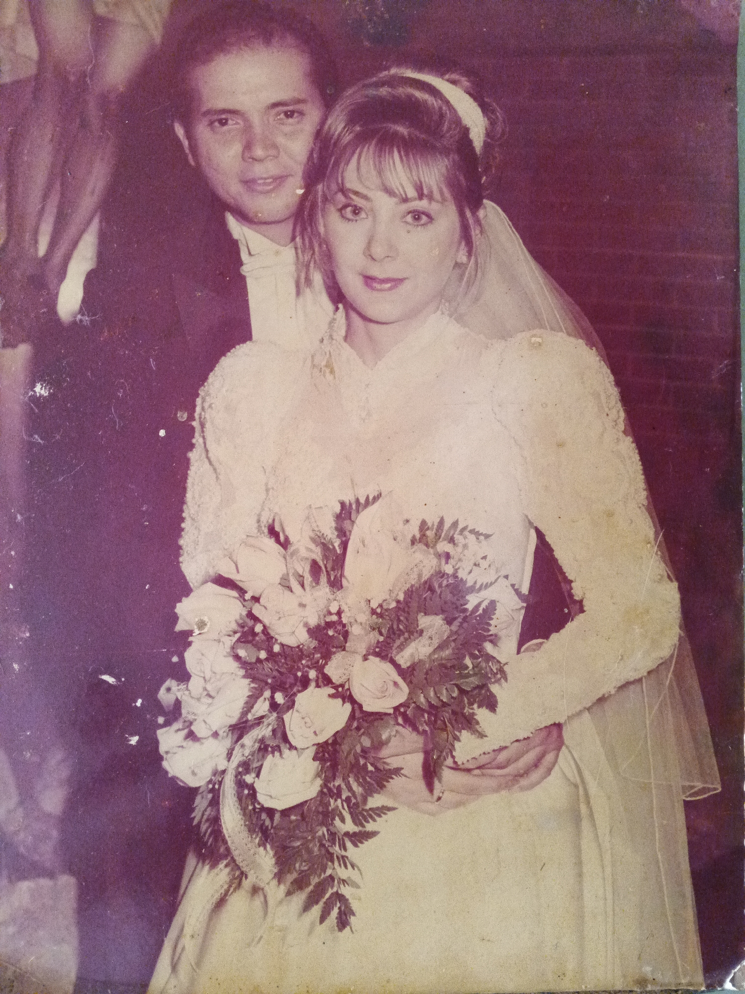 A vintage wedding photograph shows a man and woman in Caracas, Venezuela. The woman, the bride, is in the foreground, holding a bouquet of light-colored roses and ferns. She wears a long-sleeved, high-necked wedding dress with intricate lace detailing, a veil, and her hair is styled up with bangs. The man, the groom, stands slightly behind her, wearing a dark tuxedo with a white bow tie and vest. His hand is gently placed around her waist, partially obscured by the bouquet. The background is slightly blurred but suggests a brick wall or building exterior. The photo has a aged, faded appearance, with a purple hue and visible wear.