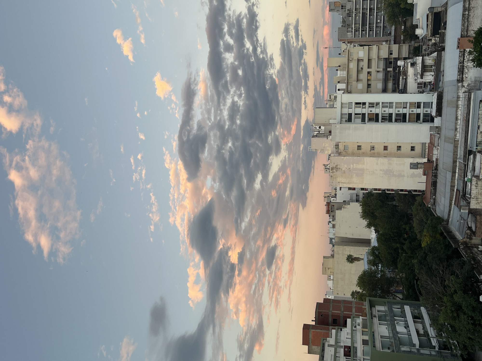 The image shows an urban scene at sunset in Cordoba, Argentina. High-rise buildings, primarily white and gray, line the right side of the frame. To the left, the sky is filled with dark, gray clouds edged with orange and pink hues from the setting sun. The light from the sunset illuminates the clouds. There are trees visible between the buildings. The scene appears to be shot from an elevated perspective, overlooking the city. No people are visible.