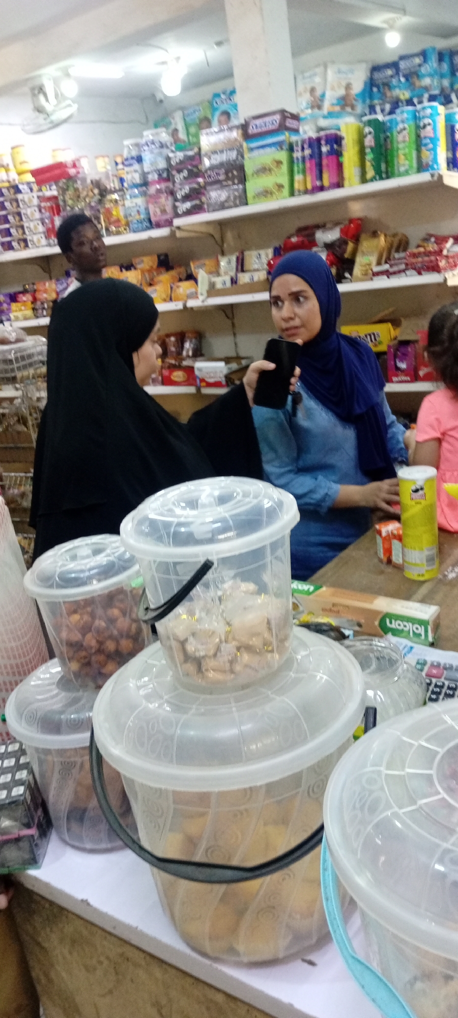 The image depicts a scene inside a store in Tudun Wada, Nigeria. Two women are engaged in conversation near a counter. One woman is wearing a black abaya, while the other wears a denim shirt and a blue hijab. The woman in the hijab is holding a black mobile phone. On the counter, several clear plastic buckets are stacked, containing what appears to be different types of snacks or food items. A yellow can of Pringles and a Falcon aluminum foil box are also visible. In the background, a dark-skinned individual stands further back in the store, near shelves stocked with various goods, including boxed items. The store appears to be well-lit with fluorescent lighting.