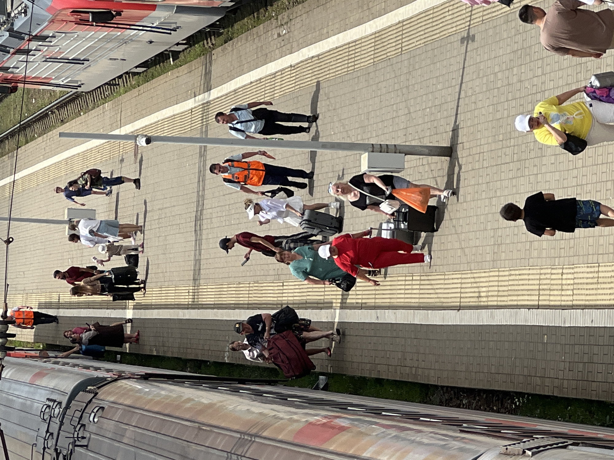 The image shows a busy train station platform in Krasnodar, Russia, with people arriving and departing. A train is parked at the platform on the left side of the image, and another train is visible on the right. Passengers are seen walking along the platform, some carrying luggage. In the center of the image, a woman in a red outfit is walking with a suitcase, and a man in an orange vest is standing, possibly a railway worker. Several other individuals are also present on the platform, some appearing to be families with children. The platform is paved with brick-like tiles, and there are yellow tactile paving strips along the edge next to the tracks. The overall scene depicts a typical day at a train station.
