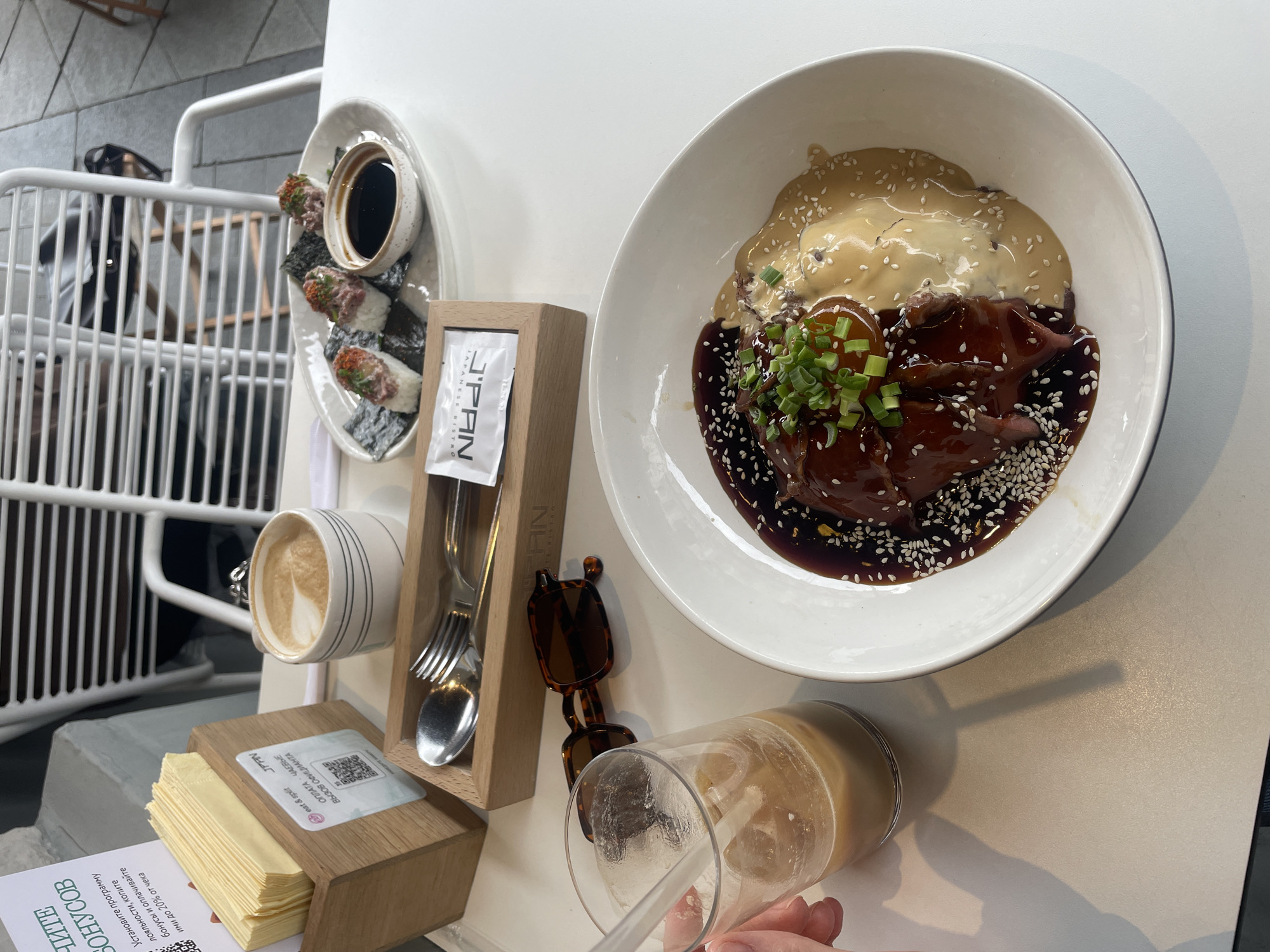 The image captures a restaurant setting. At the top right of the frame, there is a white bowl containing a dish with a shiny, dark sauce, pieces of meat, a creamy, light-colored sauce, and green garnish. To the left of the bowl, there is a wooden holder containing a fork and spoon, and a white packet with the text "J'PAN JAPANESE BISTRO" printed on it. Below the wooden holder, there is a wooden box holding folded napkins with a QR code sticker. Beside it, there is a cup of coffee with visible latte art.
On the left side of the image, sushi rolls can be seen on a small plate with a container of soy sauce. In front of the plate, there is a coffee cup with a latte on a coaster next to a rack with a white painted frame. Towards the bottom, there is a text in Russian. A pair of sunglasses are resting close to a glass with a straw, which a human hand holds. The surface the items are on appears to be a white table.
The location in the city of Unknown, Unknown, is specified by the presence of "J'PAN JAPANESE BISTRO" printed on a packet.