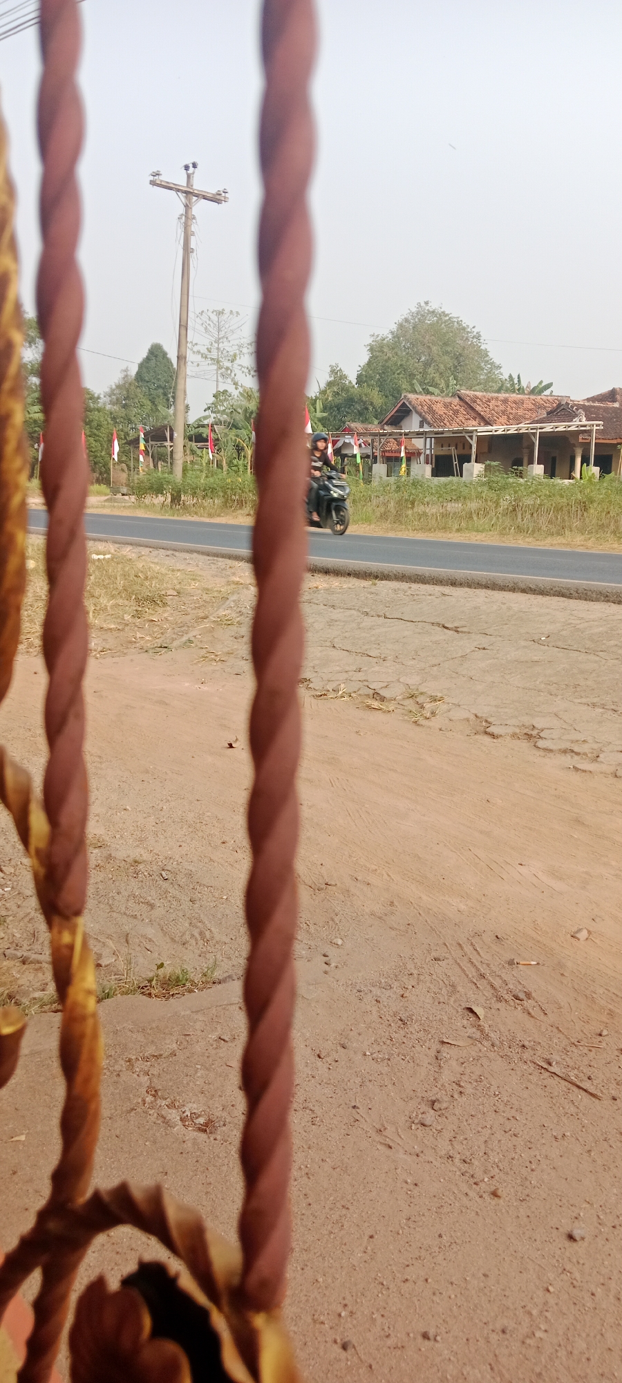 In Gondang Rejo, Indonesia, a scene unfolds featuring a road and adjacent rural landscape. The foreground is dominated by out-of-focus, twisted metal bars, one reddish-brown and the other with a metallic gold finish, creating a partial frame for the view beyond. To the left, a utility pole with ceramic insulators stands against a bright, hazy sky. Beyond the pole, the environment transitions to a paved road where a person on a black motorcycle is captured in motion, heading away from the viewer. The rider wears a dark shirt and a dark helmet. Along the roadside, several Indonesian flags on poles are visible, interspersed with lush green vegetation and some simple, open-sided structures with tiled roofs. The ground closer to the viewer is dry and sandy, with patches of cracked earth and sparse dry grass. The overall impression is one of a tranquil, everyday scene in a village or semi-rural area, possibly during daytime.