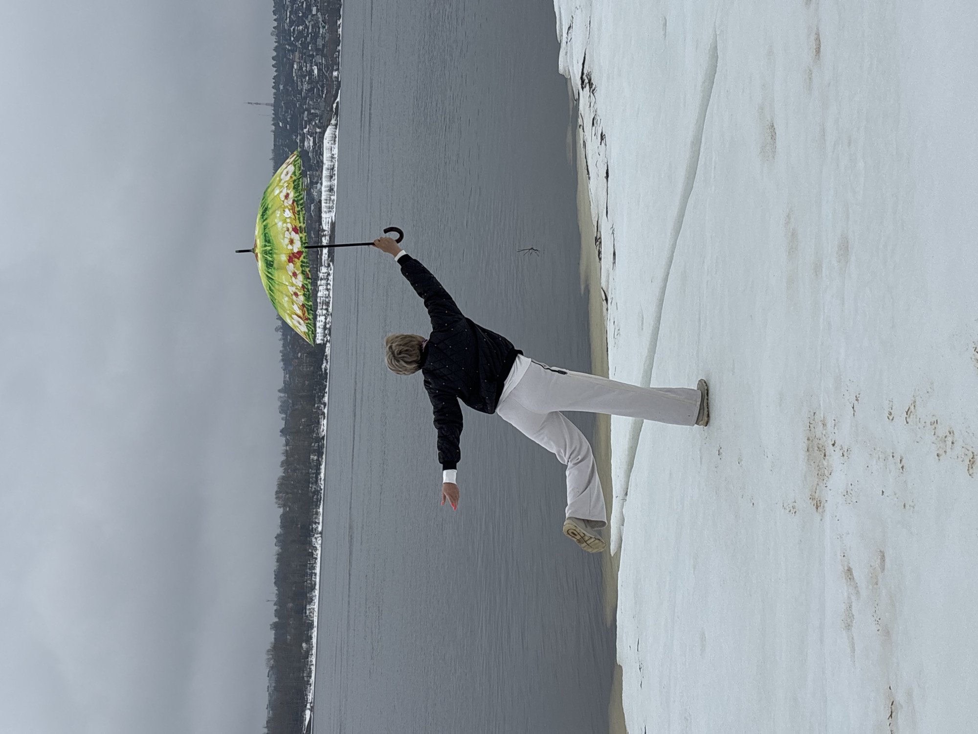 A woman stands on a snow-covered surface near a body of water. She wears white pants, a black jacket with white dots, and beige sneakers. Her left arm is outstretched towards the water, and her right hand holds a colorful umbrella overhead. The water appears calm, and a wooded area is visible across the water, indicating a shoreline. The overcast sky suggests a cold, possibly winter, day. The presence of snow indicates it is winter or early spring. The location is in the city of Chaikovsky, Russia.