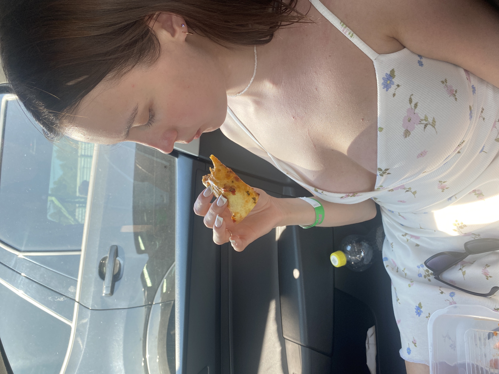A young woman is sitting in the passenger seat of a car, holding a piece of pizza to her mouth. She is wearing a white dress with a floral pattern and a thin beaded necklace. Her hair is brown and she has a green wristband on her left arm. In the background, the interior of the car is visible, including the dashboard and a bottle of water. The car appears to be parked, as suggested by the static environment and the woman's relaxed posture. It is not possible to determine the specific location within the city of Beryozovka, Russia, based on the provided image.