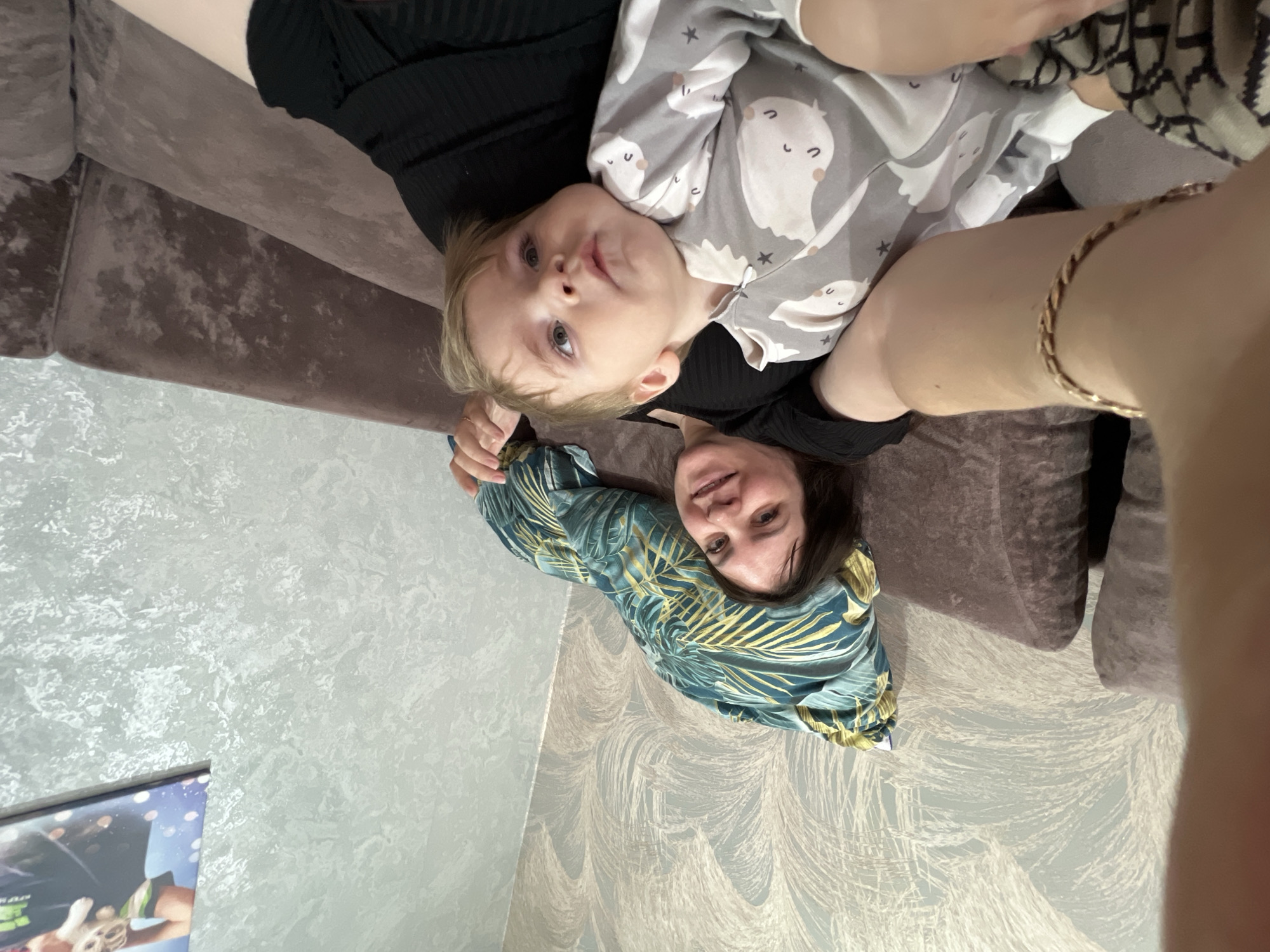 A woman is on a couch with a child in her lap. The child is wearing gray pajamas with a ghost pattern. The woman is wearing a green and yellow floral top. They are in a room with light gray patterned wallpaper. The location is implied to be in Noviy, Russia. A television screen is visible in the lower-left corner.
