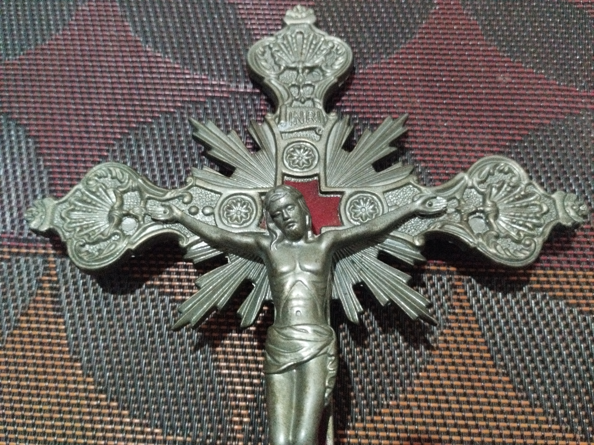 An ornate, metallic crucifix depicting Jesus Christ is positioned against a textured, woven background. The crucifix is silver-toned and features intricate details. Jesus is shown in a state of suffering, with his arms outstretched. Above his head is a plaque bearing the letters "INRI". The background appears to be a placemat or table runner with a dark, woven pattern. The lighting suggests an indoor setting. There is no discernible information in the image that places it in Sukaraja, Indonesia.