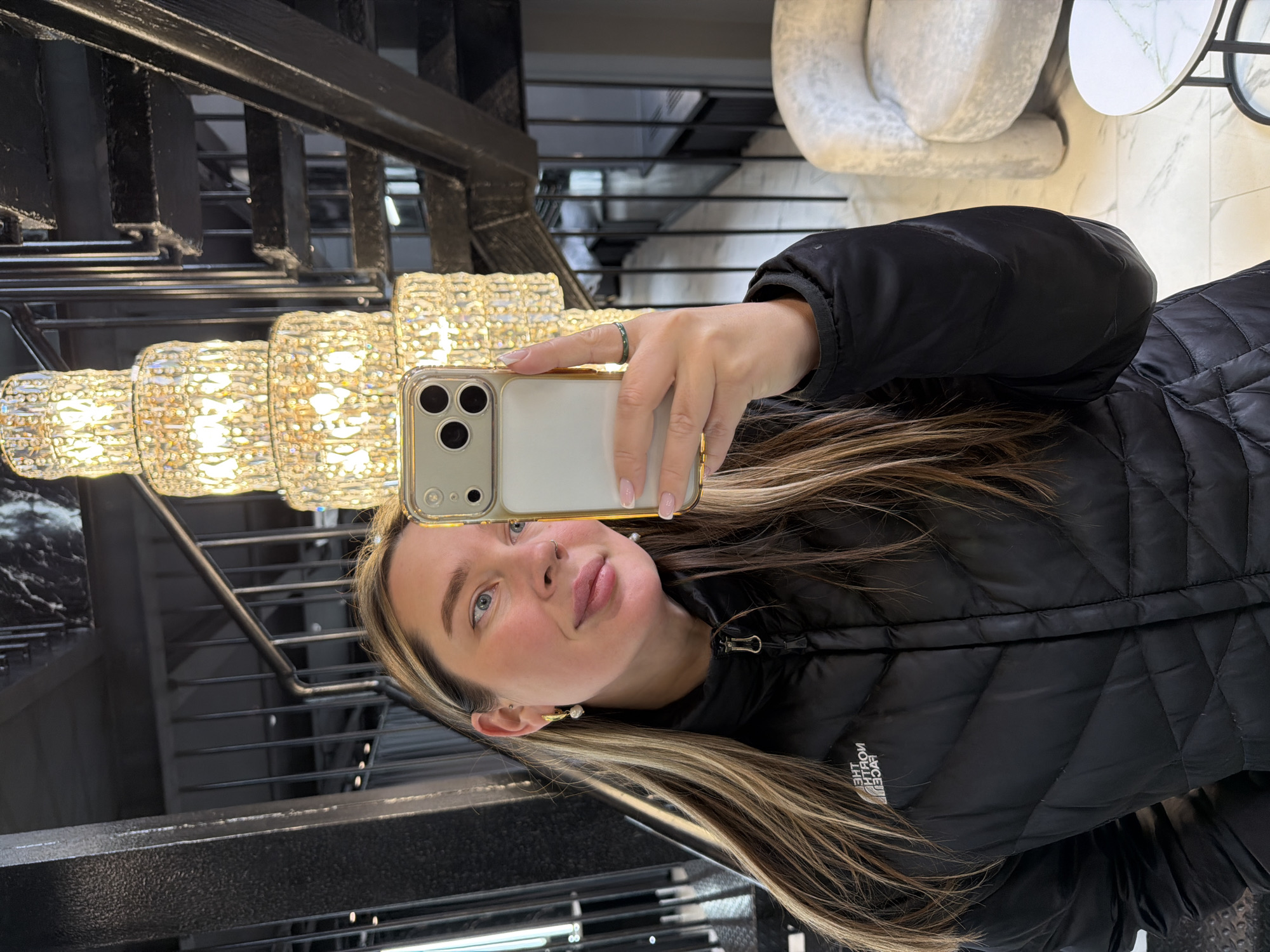 A woman is taking a selfie in a building. The location is Moscow, Russia. The woman is holding an iPhone in her right hand. She is wearing a black puffer jacket and a ring on her finger. She has long, blonde hair with highlights. In the background, there is a chandelier and a staircase with black metal railings. There is a chair and a table in the upper right corner of the image. The lighting in the building is bright.