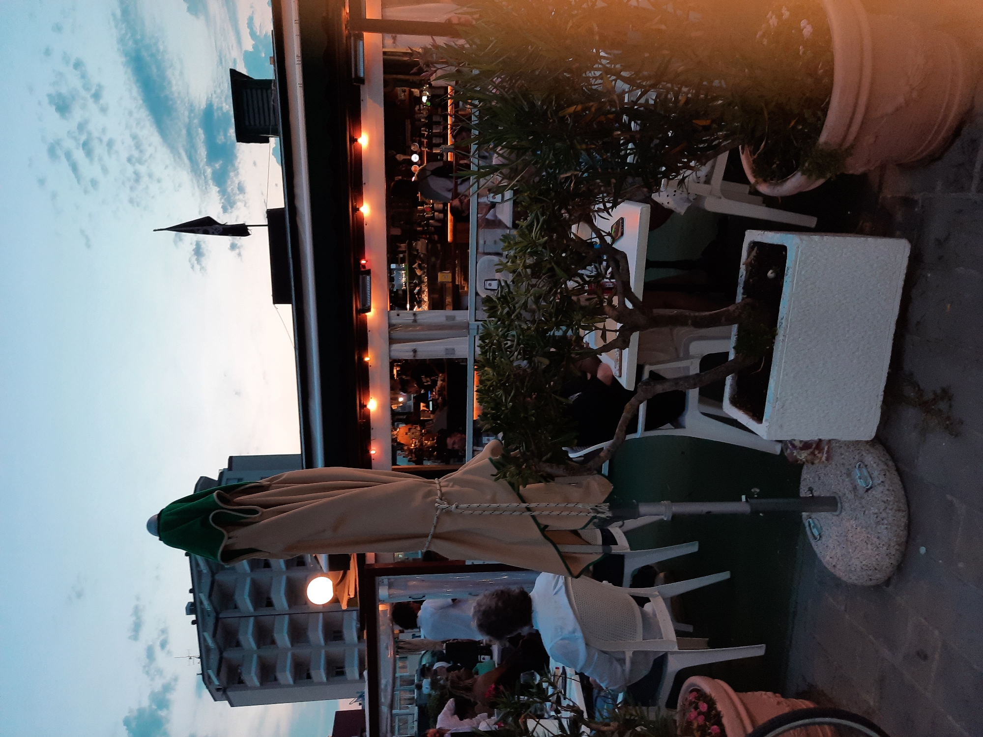 The image depicts an outdoor seating area of a restaurant or bar in Pesaro, Italy, during twilight. A large, closed umbrella with a green trim stands prominently in the foreground. White plastic chairs are arranged around tables, some occupied by patrons. In the background, a multi-story building with numerous balconies and a pirate flag flying from a rooftop are visible. The interior of the establishment shows shelves stocked with bottles and lit by warm lighting, with several people working or socializing behind the counter. The overall scene suggests a relaxed evening atmosphere at a coastal establishment.