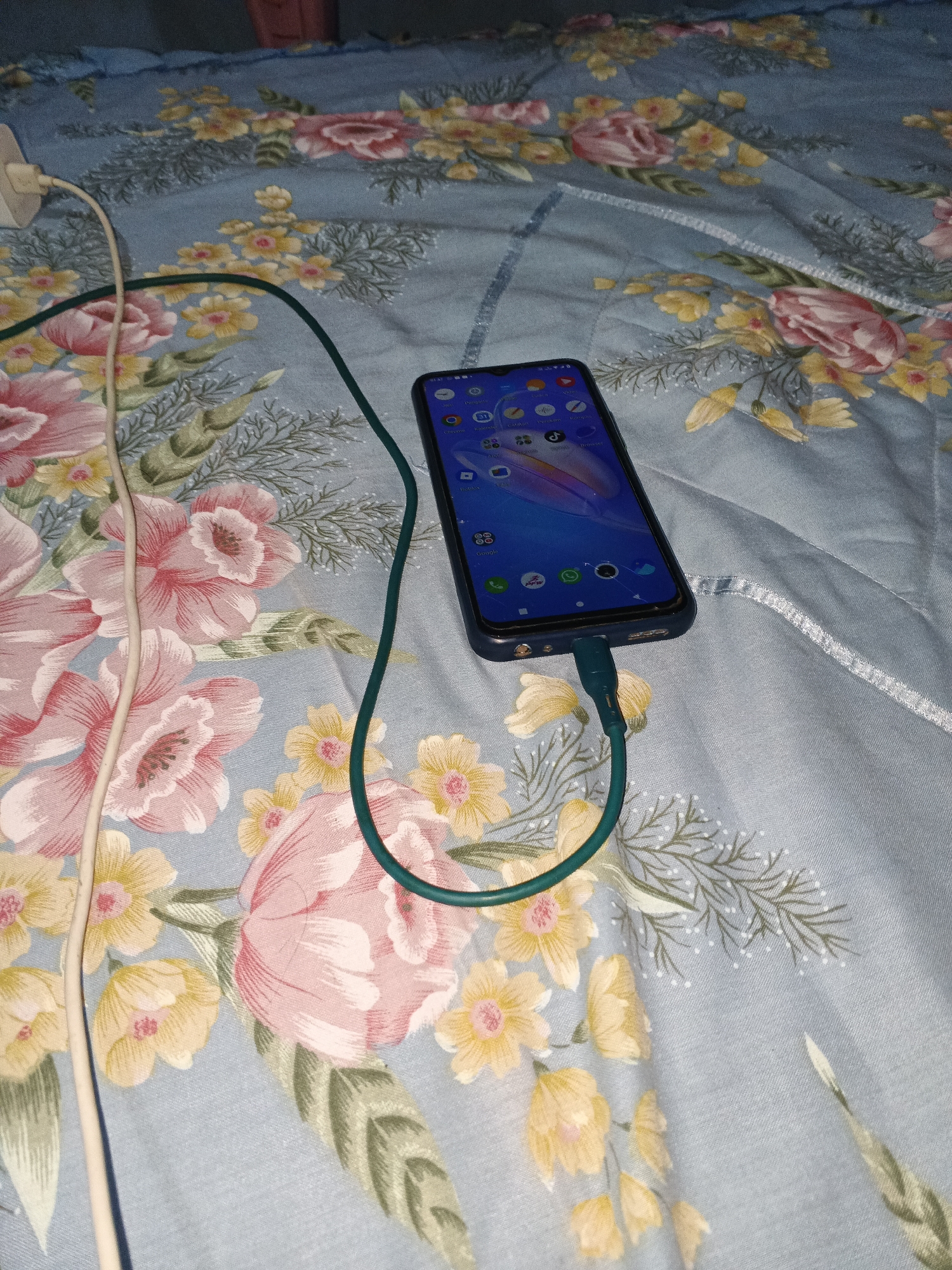 The image shows a smartphone, charging cable, and charger on a floral patterned blue bedsheet. The smartphone is connected to the green charging cable, which is then connected to a white charger. The charger is plugged into an electrical outlet that is partially visible. The bedsheet has a floral pattern with pink, yellow, and green flowers. The image is taken from a top-down angle, with the phone in the center of the frame. The scene is indoors, likely in a bedroom. There are no people visible in the image. The overall impression is one of domesticity and everyday life. The image is not taken in Warungkiara, Indonesia, but rather in a domestic setting.