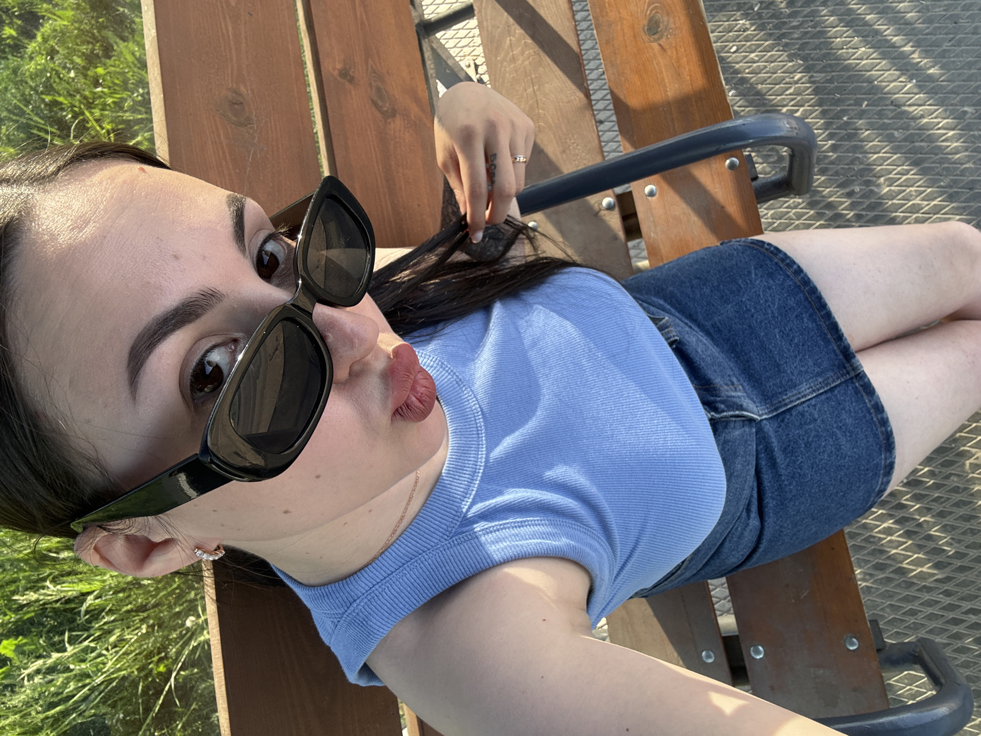 A woman is lying on a wooden bench. She is wearing sunglasses, a blue shirt, and a denim skirt. She has a ring on her finger. The bench has a metal grid underneath and is located outdoors in a sunny environment. The location is Chaikovsky, Russia.