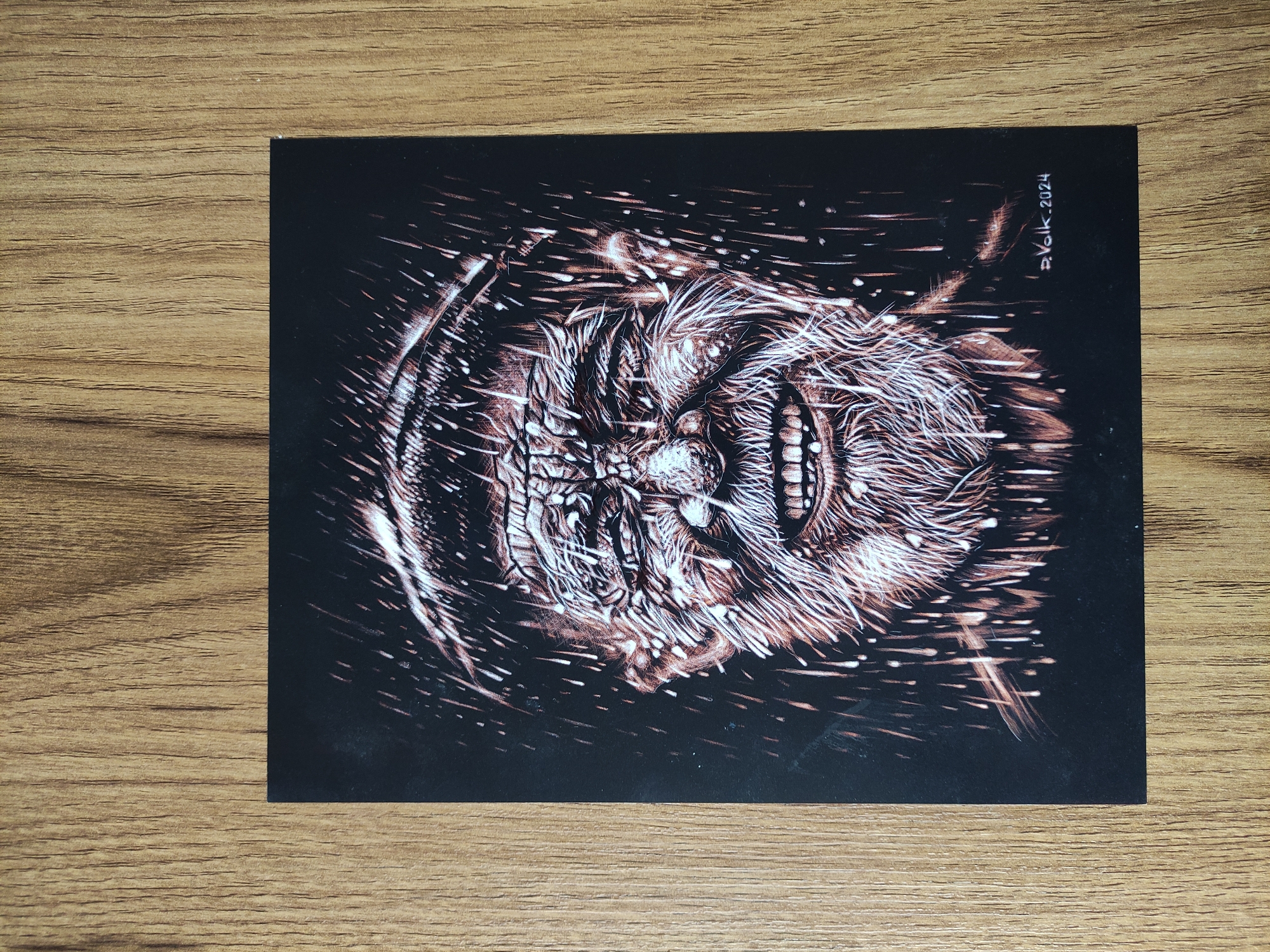 The image shows a postcard with a black background featuring an illustration of a face with prominent teeth. The drawing style uses brown and white lines to create a textured, somewhat grotesque appearance. The name "D. Volk 2024" is written in the upper right corner. The postcard is placed on a wooden surface with a visible grain pattern. The location and presence of people or interactions are not discernible. The image is forensic and matter-of-fact in that it provides a clear and unemotional depiction of the scene and the visible details of the objects present.