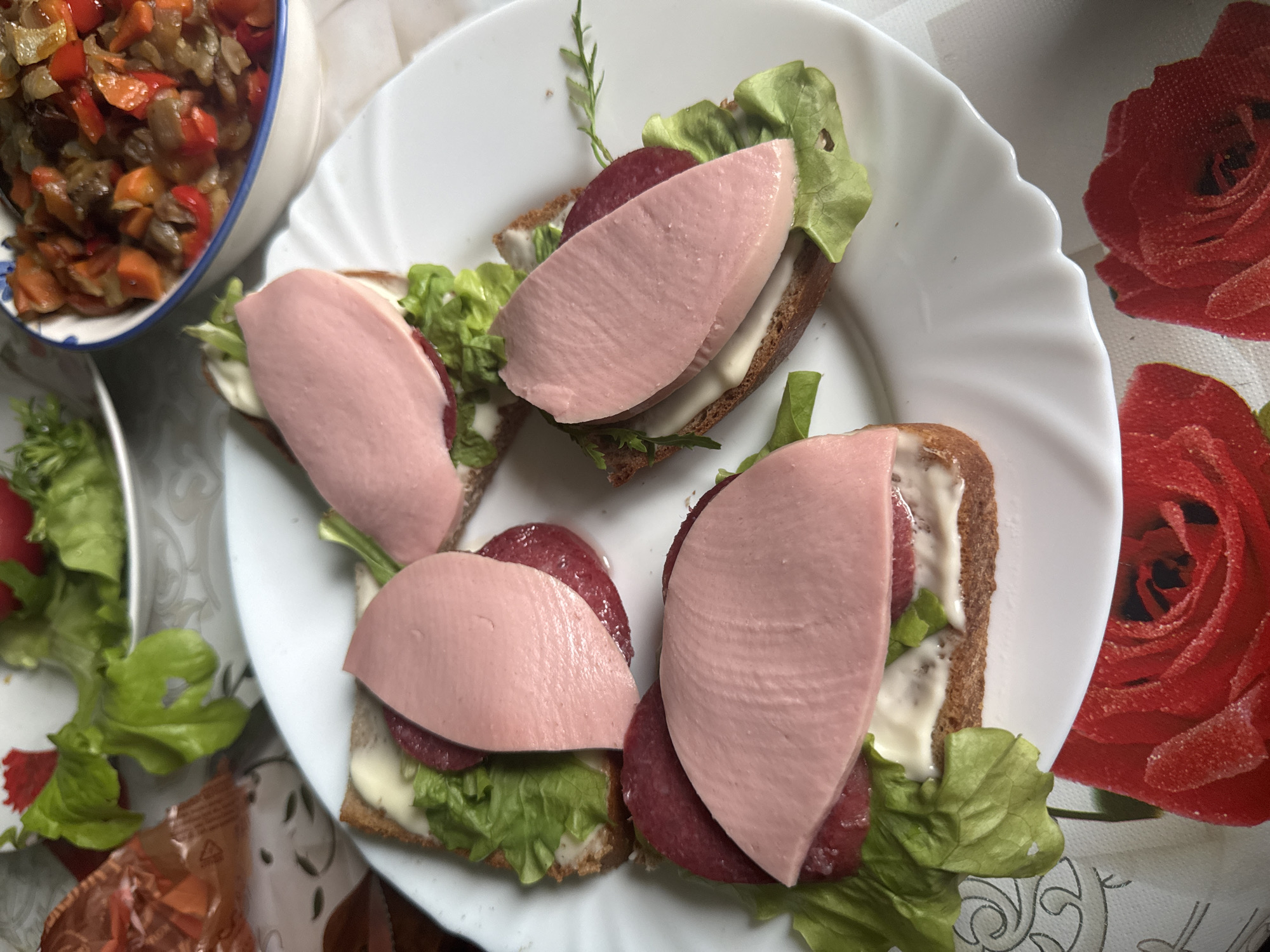 Four open-faced sandwiches are arranged on a white, fluted plate. Each sandwich consists of a slice of dark bread topped with mayonnaise, a lettuce leaf, a slice of salami, and a thick slice of pink, processed meat, possibly bologna. In the background, a bowl of mixed vegetable stew or ratatouille is visible, along with a bowl of lettuce. The scene is set on a tablecloth with a red rose pattern. The image appears to be taken indoors. The prompt mentions the city of Красное, Russia, but there is no visual information in the image to confirm or deny this specific location; the scene is generic and could be anywhere.