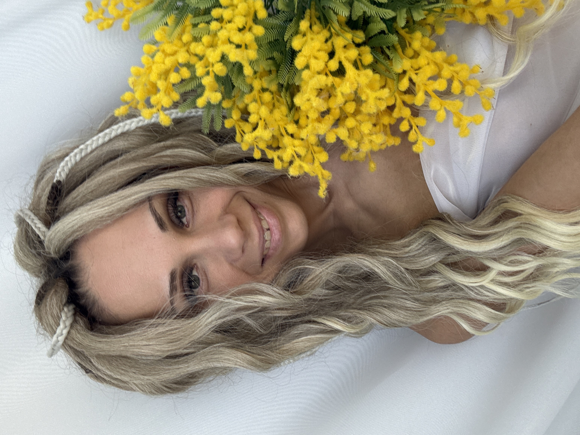A woman lies on a white sheet with yellow mimosa flowers arranged near her head. Her hair is blonde with braids near her forehead. The location is not evident from the image.