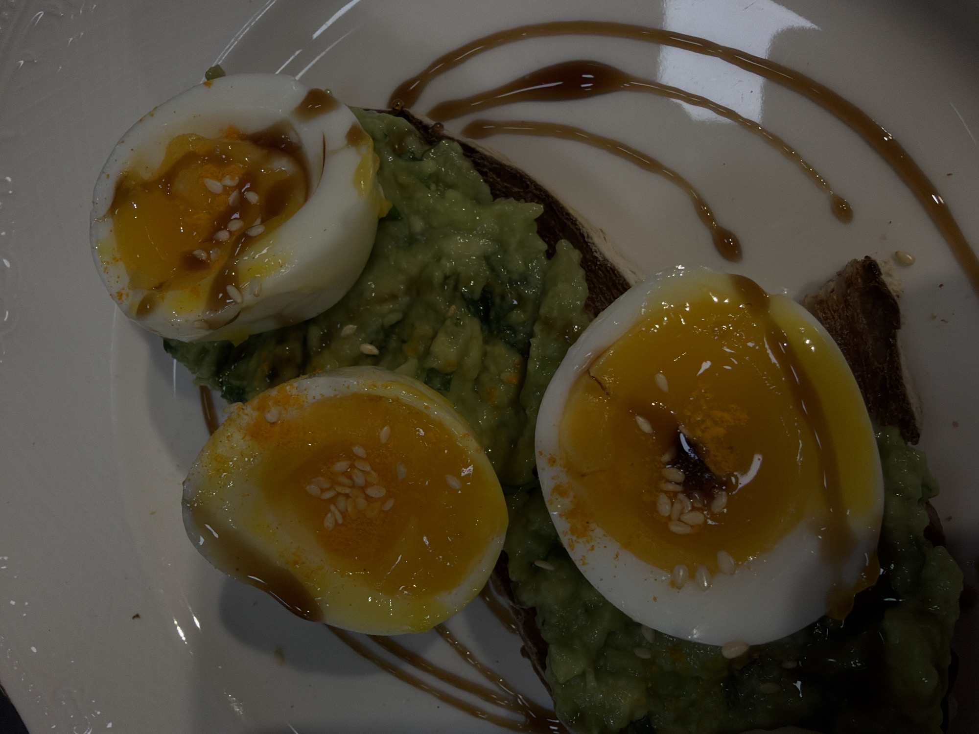 The image shows a close-up of a plate of avocado toast topped with sliced soft-boiled eggs, garnished with sesame seeds and a drizzle of sauce. The dish is served on a white plate. The background is slightly blurred, indicating a shallow depth of field. The location and city of Yerevan, Armenia cannot be determined from the provided image.