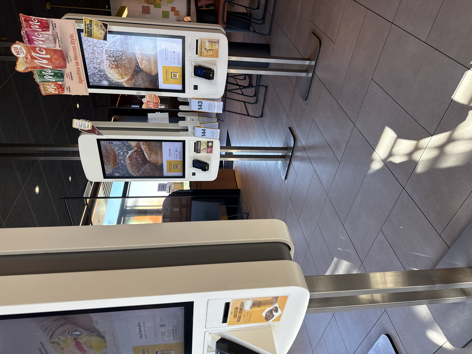 The image shows an interior view of a McDonald's restaurant in Werther, Germany, likely a self-service ordering area. In the foreground, three digital ordering kiosks are visible, two in sharper focus. The kiosks have large touchscreens displaying food items, with payment card readers and slots for cash or coupons below. One kiosk has a promotional flyer with an image of McCafe coffee and cookies. The floor is tiled with large square tiles. In the background, there are tables and chairs, suggesting a dining area. The lighting appears to be a mix of artificial ceiling lights and natural light coming from windows, casting shadows on the floor. The overall environment is clean and modern, typical of a fast-food establishment.