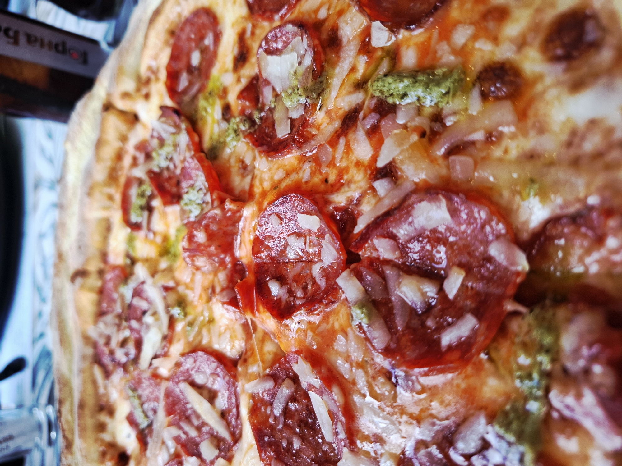 The image shows a close-up of a pizza topped with pepperoni, shredded cheese, and small dollops of pesto. The crust appears golden brown. The location and specific city are unknown.