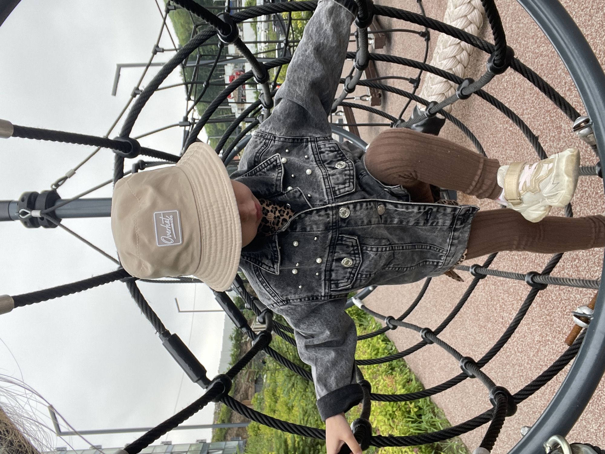 A young child, likely a girl, is climbing a black rope jungle gym structure. She is wearing a tan bucket hat with a white embroidered logo, a gray denim jacket adorned with pearl-like studs, a leopard print top visible at the collar, brown ribbed leggings, and light-colored chunky sneakers. The child's right leg is bent and placed on one of the ropes, while her left hand is grasping another rope for support. The jungle gym is set outdoors, with a grey, overcast sky above. In the background, there is lush green foliage and some urban structures, including modern buildings. A paved pathway with a reddish-brown speckled surface is visible below the jungle gym. The overall setting suggests a park or a children's play area within a city.