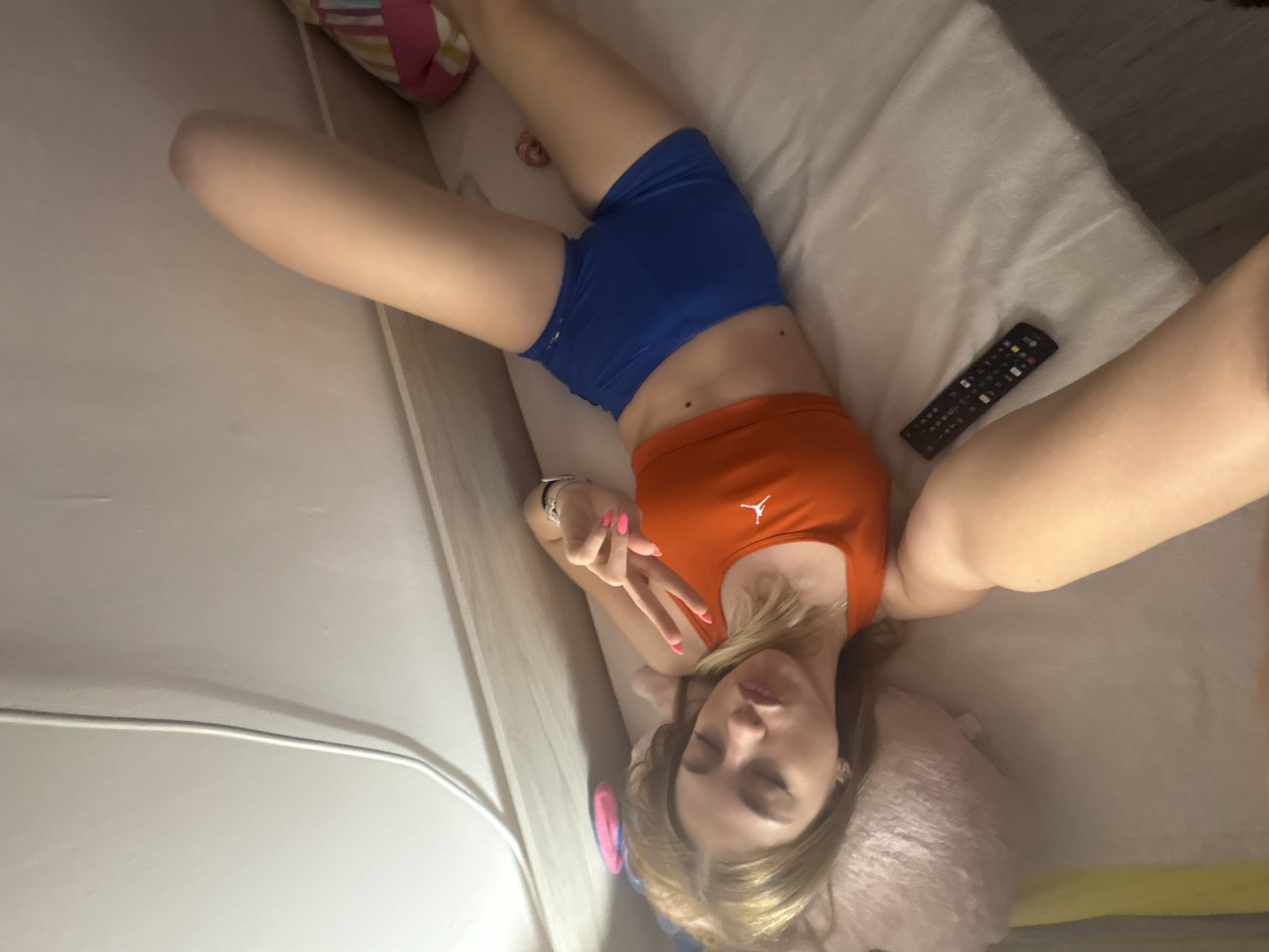 A young woman is lying on her back on a light-colored bed or couch, viewed from a top-down, rotated perspective. She is wearing an orange crop top with a white "Jumpman" (Jordan) logo on the chest, and blue shorts. Her legs are extended, and her arms are resting by her sides. With her left hand, she makes a peace sign, prominently displaying pink nail polish on her fingers and a silver-toned watch on her wrist. Her eyes are closed or partially closed, and her blonde or light brown hair is spread around her head, which rests on a light pink, fluffy pillow. Two small moles are visible on her abdomen. To her right, a black remote control is resting on the bedding. In the upper left background, a multicolored striped pillow is partially visible. A white cable runs along a light-colored wall in the lower part of the frame. The setting appears to be indoors with soft, ambient lighting, conveying a relaxed atmosphere.