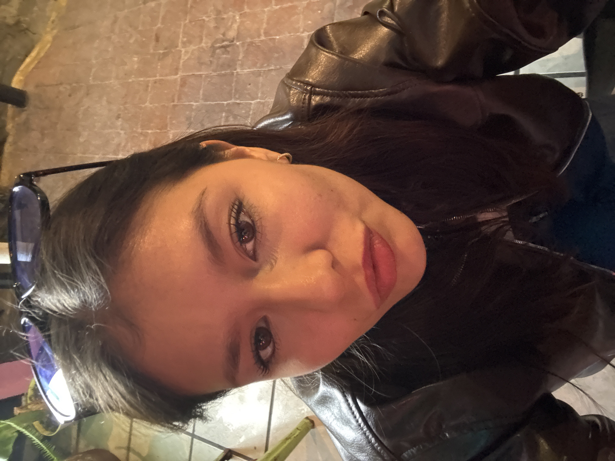 A young woman poses indoors, wearing a brown leather jacket. A pair of eyeglasses rests near her on a table. The background includes a floor with brick-like patterns and a glimpse of green foliage. The image was taken in San Andrés Cholula, Mexico.
