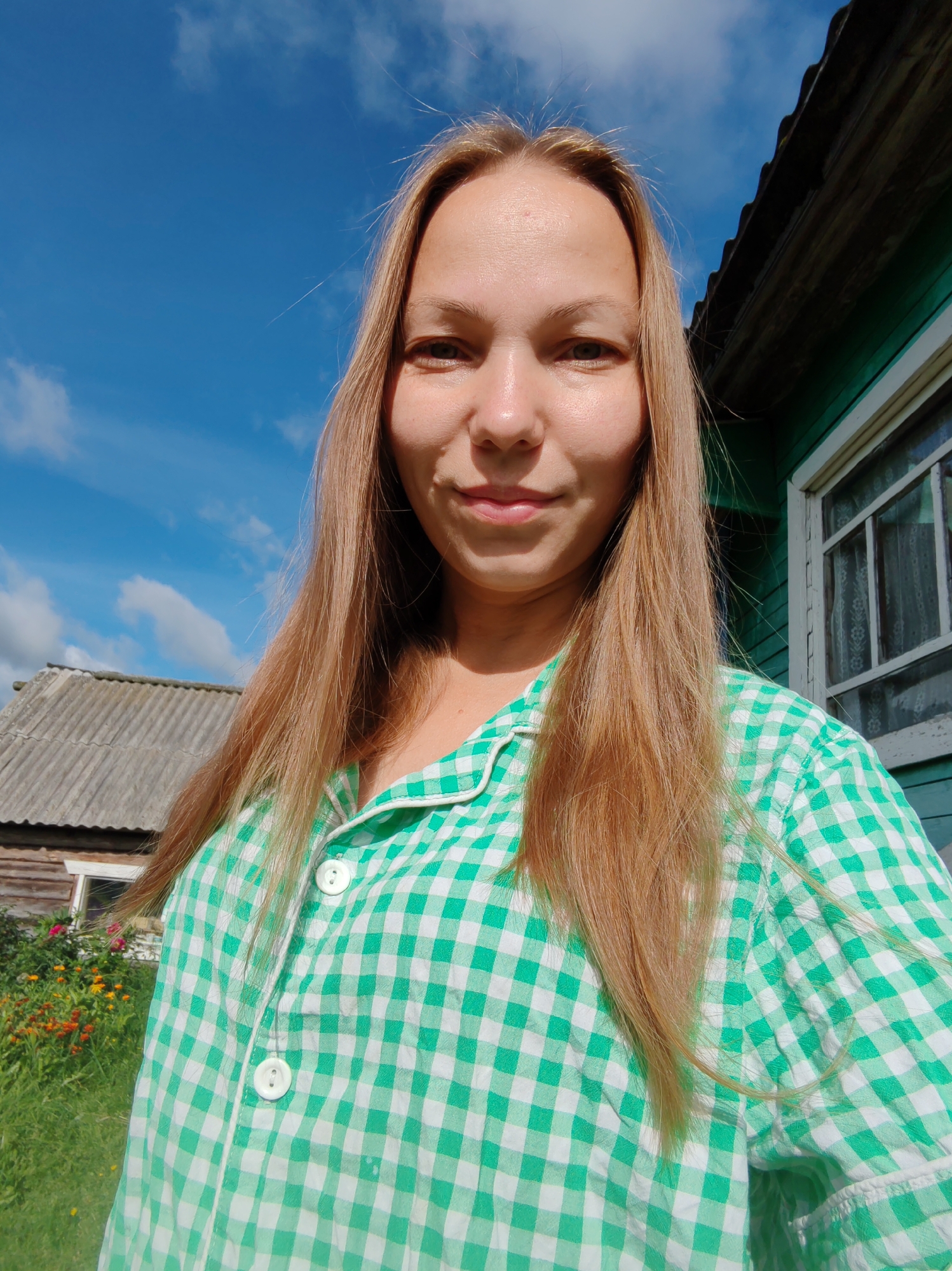 A medium-shot selfie of a young woman with long, blonde hair is taken outdoors, with a bright blue sky and scattered clouds in the background. To her right, a weathered green wooden building with a window is visible, and to her left, a rustic-looking grey-roofed structure and a patch of colorful flowers in a garden are present. The woman is wearing a green and white checkered shirt. The scene suggests a rural or suburban setting, possibly in a village or town in Russia, given the architectural styles and natural elements. The lighting indicates it is daytime, with sunlight illuminating the scene.