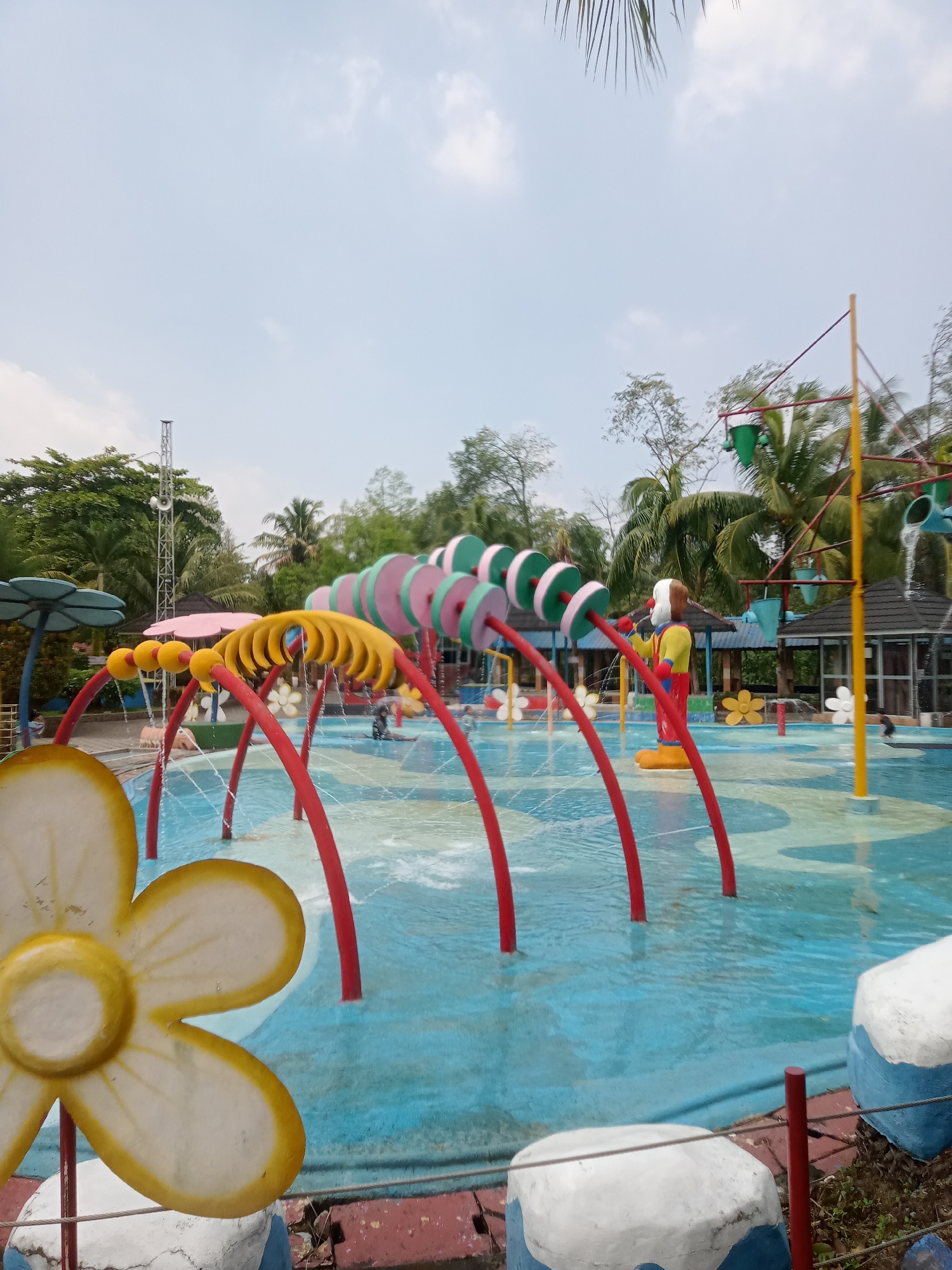 This image depicts a shallow water park, possibly a playground within a larger park or resort, in Baru, Indonesia. The foreground features large, stylized flower decorations with yellow centers and white petals, one of which is prominently positioned on the left side of the frame. The water park itself is filled with blue water and has a multi-colored, patterned floor. Several water features are active, with streams of water spraying from red, arching structures.
In the midground, there's a clown-like figure standing in the water, wearing a yellow and red outfit. Behind the clown, a series of colorful, circular structures are visible, also contributing to the water play. Further in the background, there are trees and foliage, indicating an outdoor setting. A tall, metal tower with speakers is visible on the left side of the image, and a small, dark-roofed building is present on the right. The sky above is partly cloudy. The overall scene suggests a family-friendly recreational area.