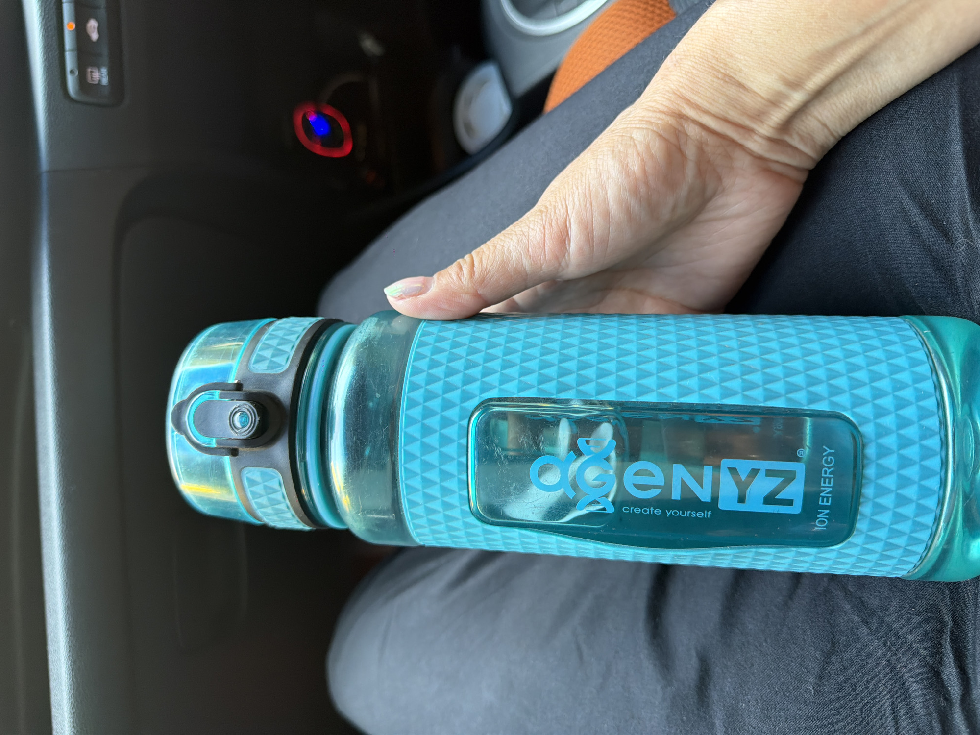 A person's hand holds a turquoise water bottle inside a vehicle. The bottle has a textured grip and a clear window with the brand "AGENYZ" and the text "create yourself" and "ION ENERGY" visible. The interior of the car is dark, with hints of car controls and dashboard lighting visible in the background. The scene is set in a vehicle, not in the city of Turka, Russia.