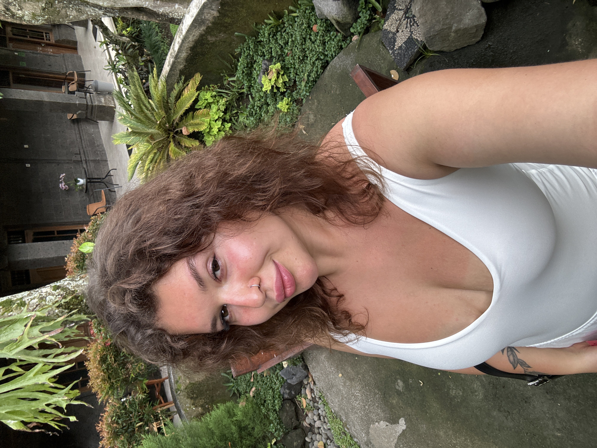 This close-up outdoor selfie captures a young woman with light skin and brown, curly hair looking directly at the camera with a soft, pleasant smile. She has a silver hoop piercing on her right nostril and a visible tattoo featuring a leaf or branch design on her right bicep/tricep area. She is wearing a white, low-cut tank top or dress.
The setting appears to be a lush tropical garden or resort courtyard, consistent with the Ubud District, Indonesia context. The background features dense green foliage, including ferns, ground cover plants, and a prominent cycad-like plant. There are also grey concrete or stone structures, possibly pathways, planters, and the edge of a water feature, along with scattered dark rocks and small pebbles. In the further background, multi-story buildings with balconies or patios are visible, adorned with small tables and chairs, suggesting accommodation. The lighting is soft and diffused, indicating daytime, possibly overcast conditions or shade. No visible text is present in the image. The overall mood is relaxed and natural.