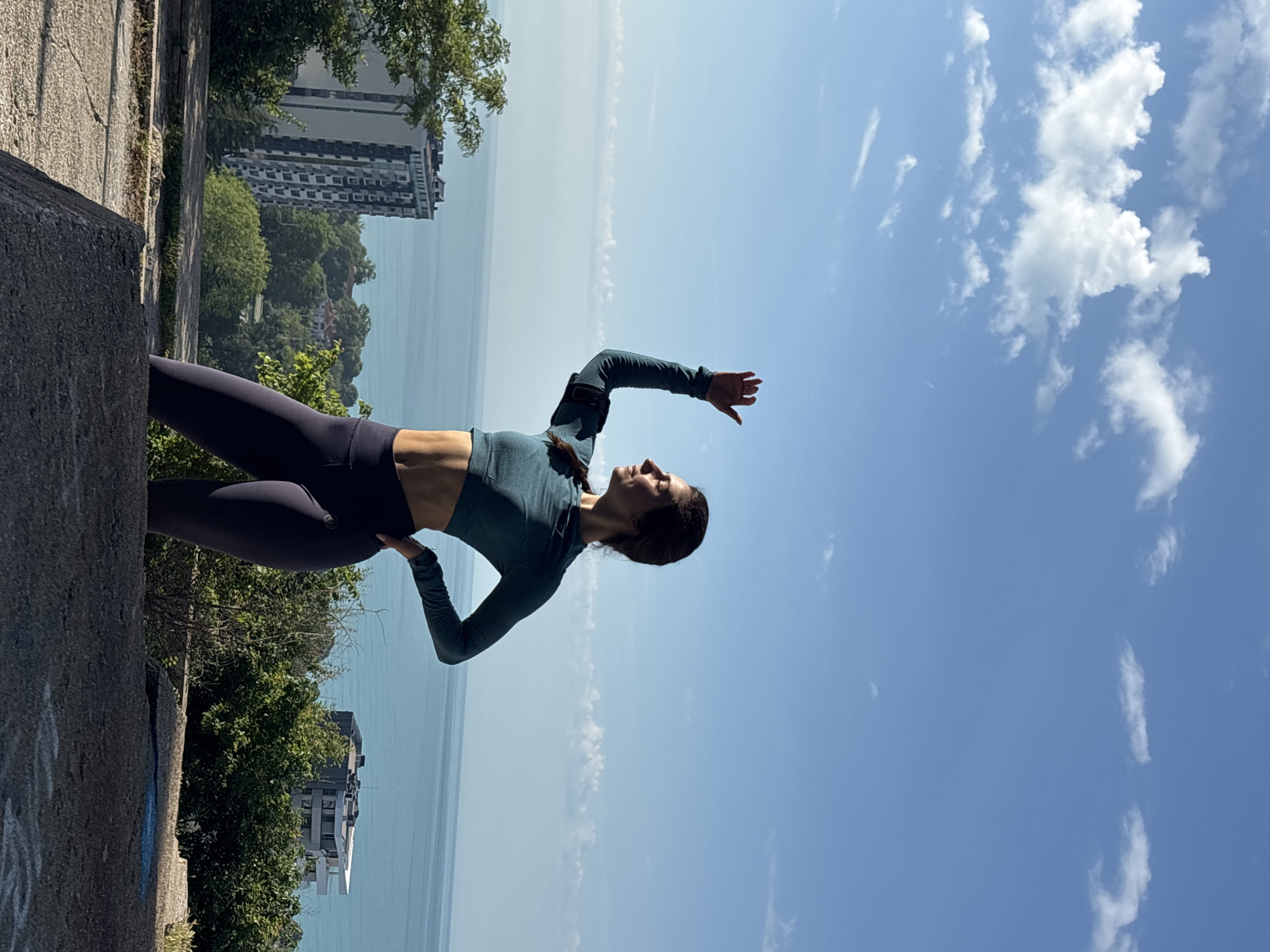 A full-shot outdoor photograph, correctly oriented, depicts a woman striking an athletic or yoga pose against a scenic backdrop. The woman, appearing to be in her 20s or 30s with dark hair, wears a long-sleeved teal crop top and dark athletic leggings. She is captured in profile, looking upwards with a serene expression, her right arm extended towards the sky and her left arm bent at her side. A small black strap is visible on her right upper arm, possibly a fitness tracker or brace.
She stands on a rugged concrete ledge or wall, which occupies the lower left portion of the frame. Beyond the ledge, a vibrant green landscape with numerous trees and foliage extends towards a wide expanse of calm, blue sea, characteristic of the Black Sea in Varna, Bulgaria. In the distance, a city skyline with several buildings, including one prominent taller structure, is visible on the horizon. The sky above is a brilliant clear blue, dotted with scattered white cumulus clouds, indicating a bright and sunny daytime. The overall atmosphere conveys a sense of tranquility and focus. There is no visible text in the image.