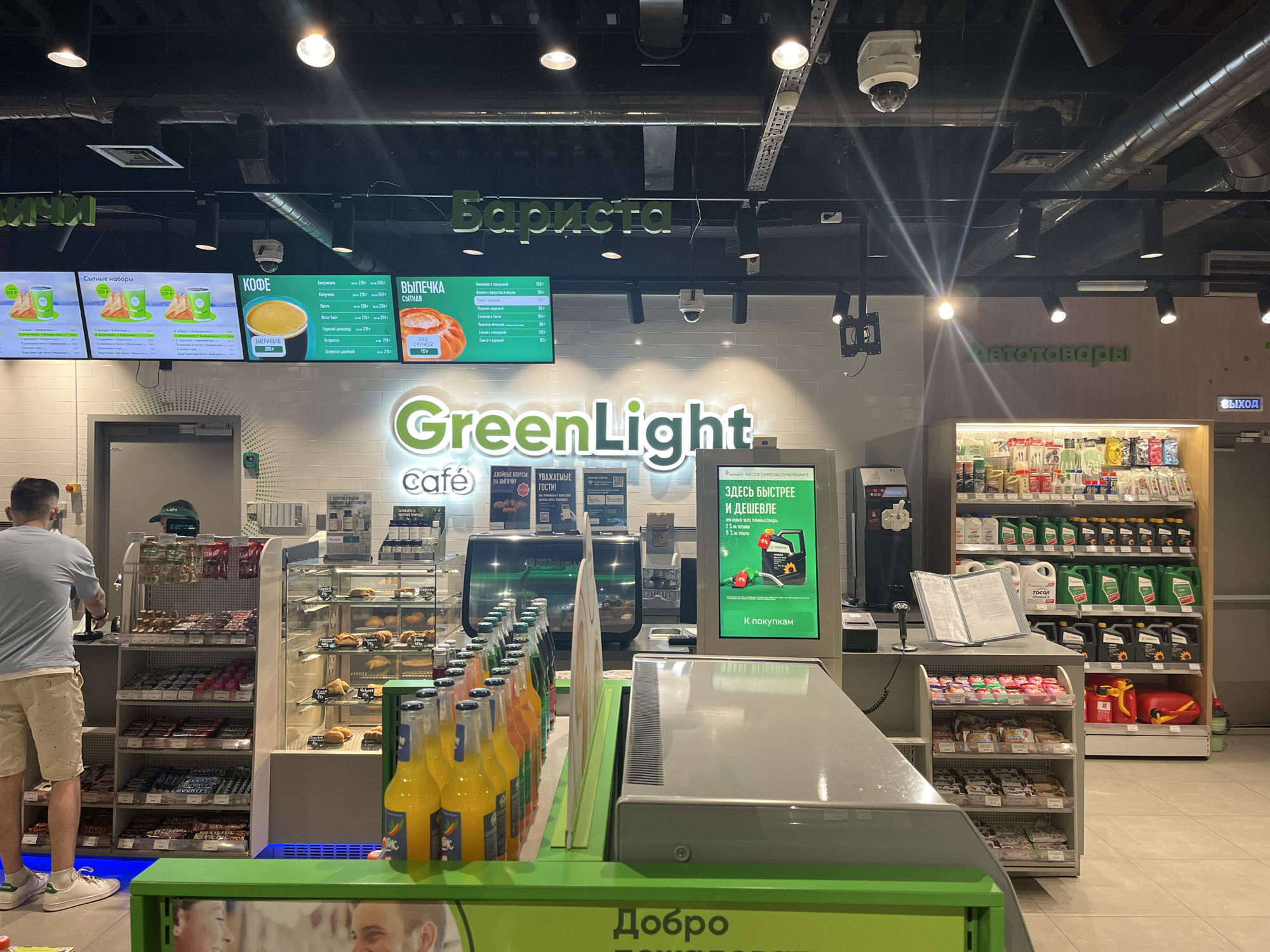 The image shows the interior of a GreenLight Café in Kazan, Russia. A man in a light blue shirt and khaki shorts stands on the left, facing the bakery section. A counter with rows of bottled drinks is in the foreground. A self-checkout kiosk is to the right. Above the counter, a menu board lists various items in Russian, including sandwiches, coffee, and pastries, along with their prices. There are shelves stocked with various automotive products, including motor oil. The store signage includes the café name, sections labeled "Barista" and "Avtotovary" (auto goods), and promotional materials for discounts and offers. The cafe's design appears modern and well-lit.